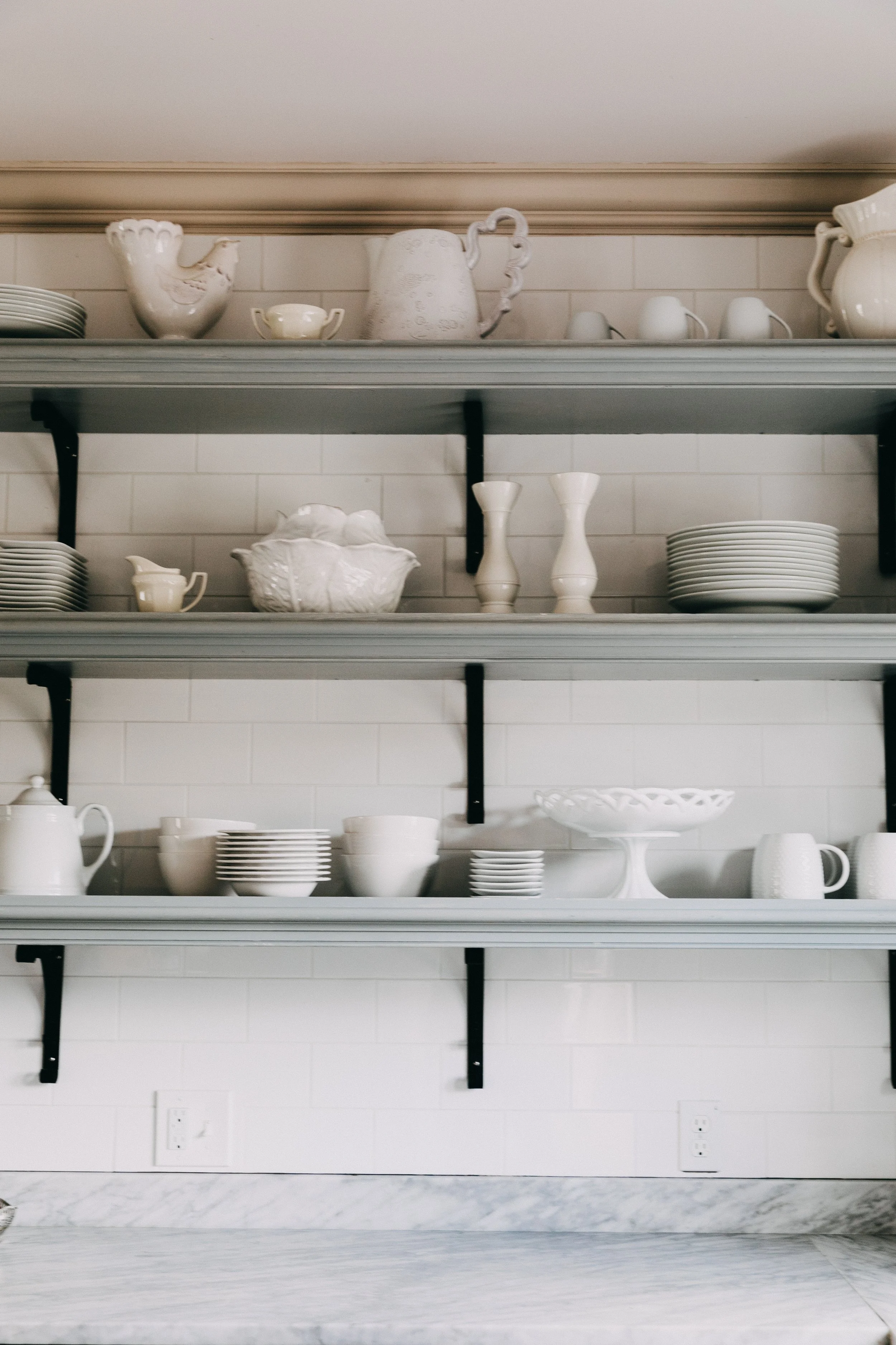 Shelves filled with white dishes, bowls, cups, and teapots against a white brick wall.