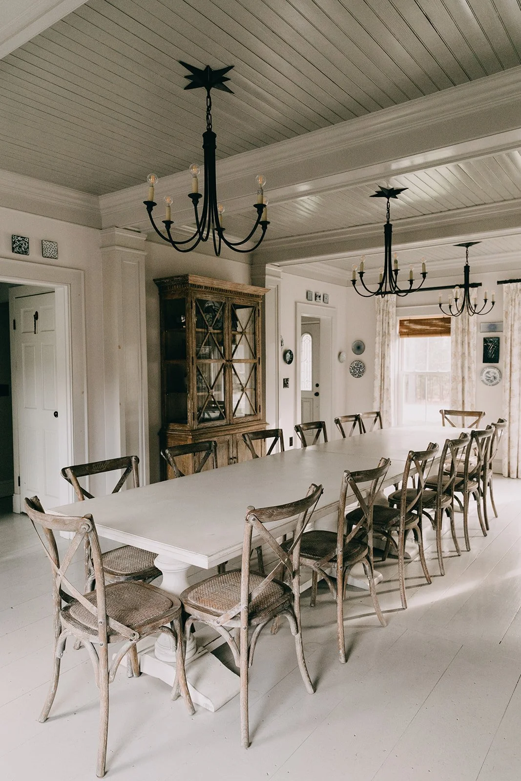 A spacious dining room with a large white table and multiple wooden chairs. There are three black chandeliers hanging from the ceiling, and a wooden cabinet against the wall. The room has a large window with sheer curtains, allowing natural light to 