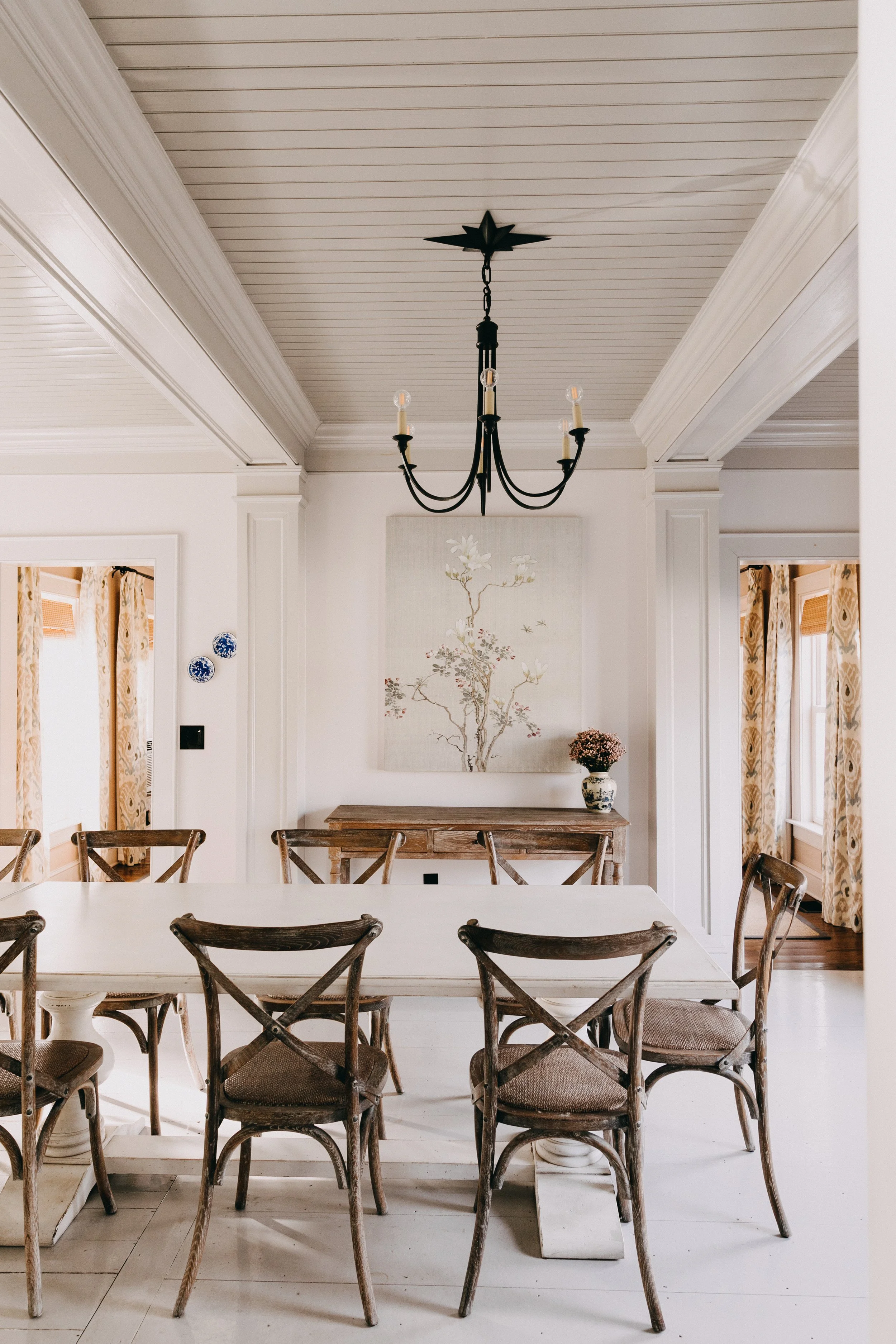 A dining room with a white table, wooden chairs, a black chandelier, a painting of a tree on the wall, and a vase of flowers on a wooden sideboard. Curtains with yellow and beige patterns are on the windows.