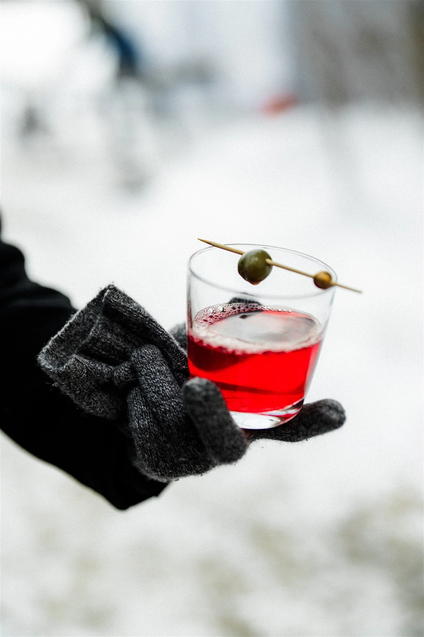 A person wearing gray gloves holding a clear glass with a red beverage, garnished with a skewer of green olives, in an outdoor winter setting with snow.