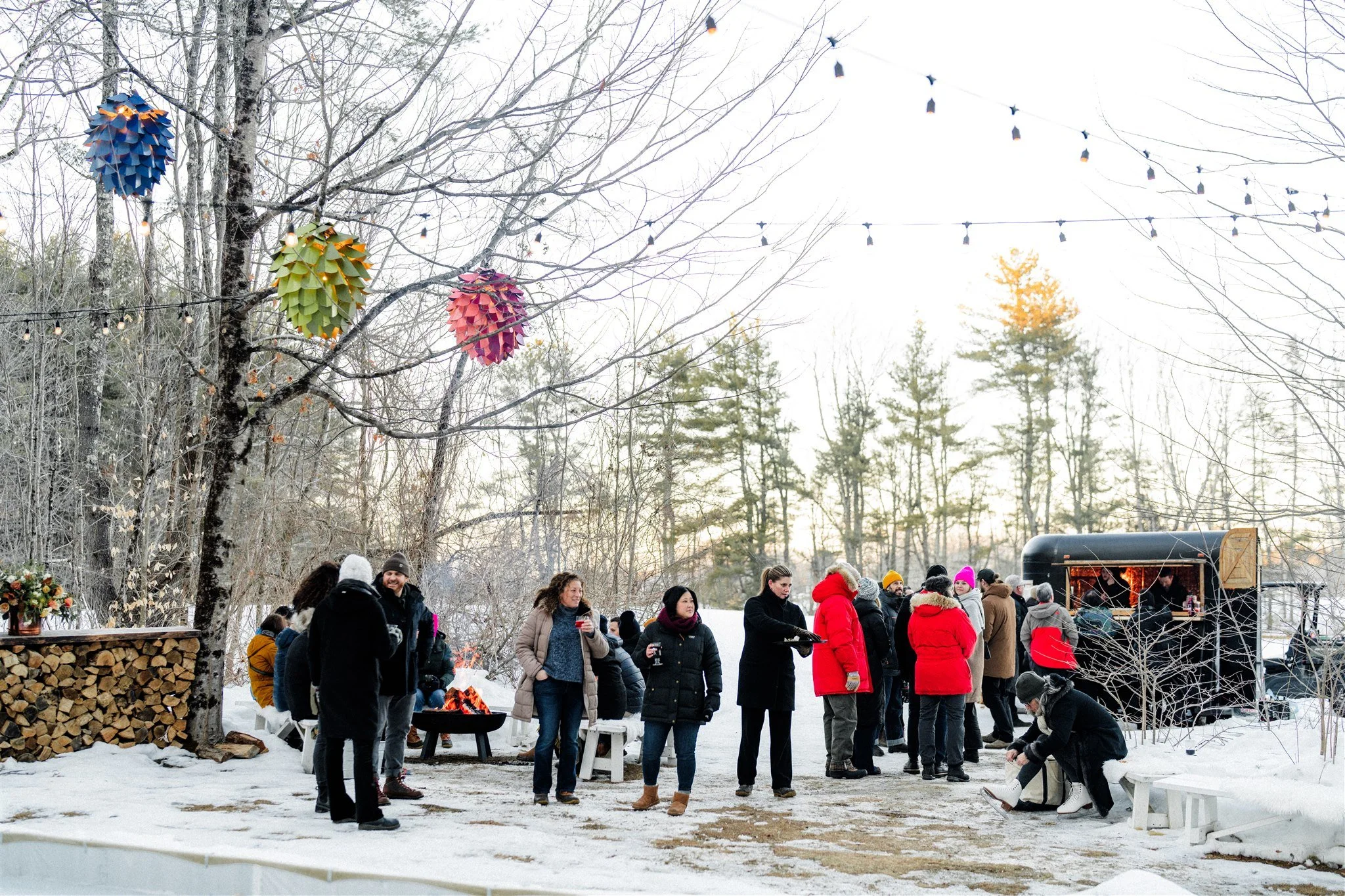 group of people gathered outdoors in a snowy setting, some standing and talking, others sitting near a fire pit and food truck, with colorful paper lantern decorations and string lights hanging from bare trees.