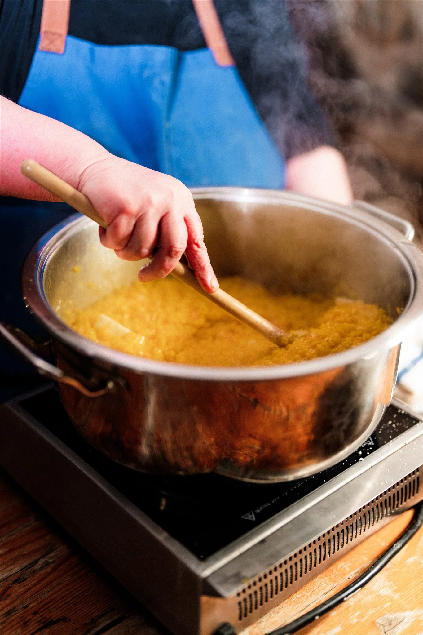 A person stirring a pot of orange lentil soup on an electric stove, wearing a blue apron.
