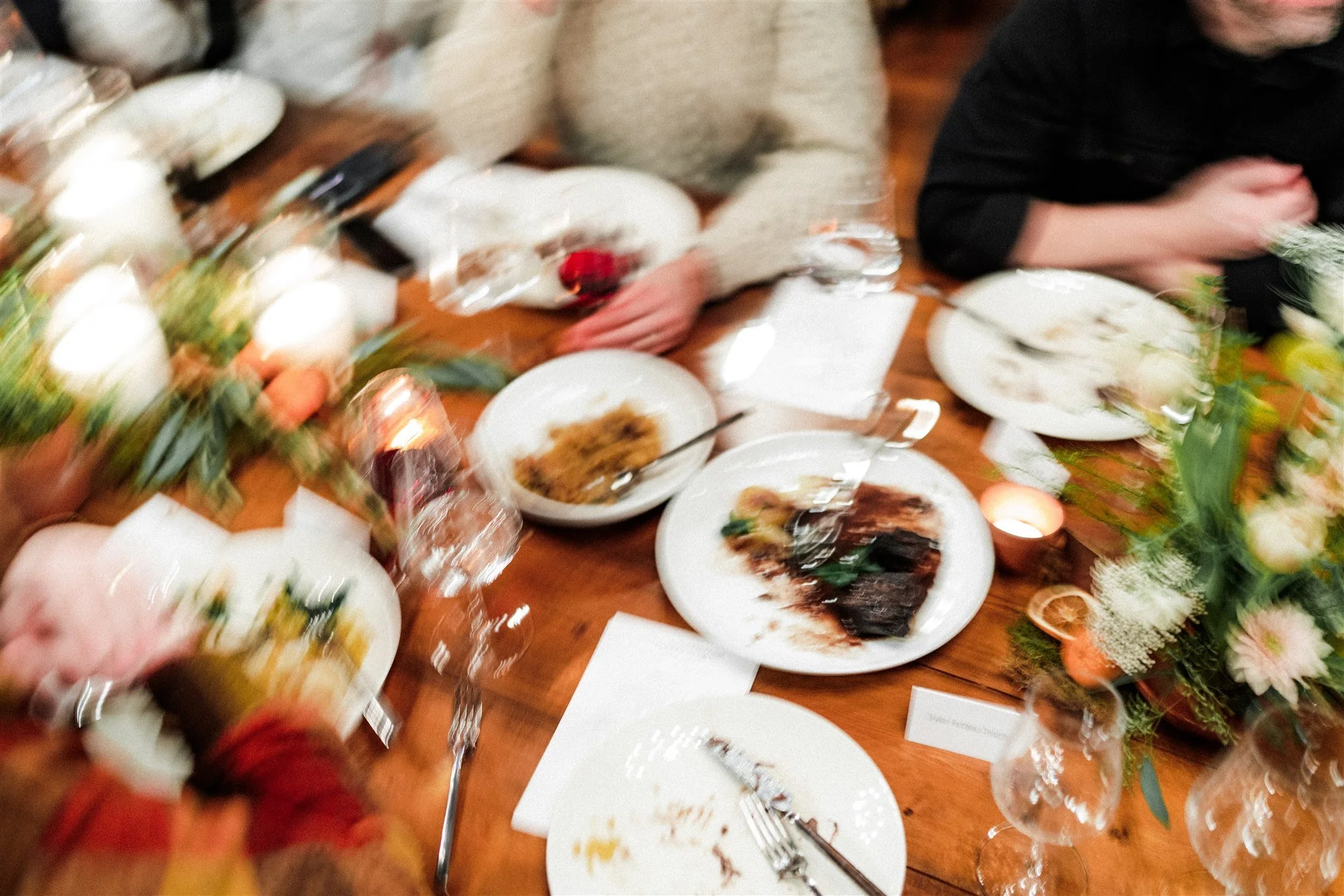 A blurry photo of a dining table with plates of food, flowers, candles, and glasses, with people seated around it, celebrating a meal.