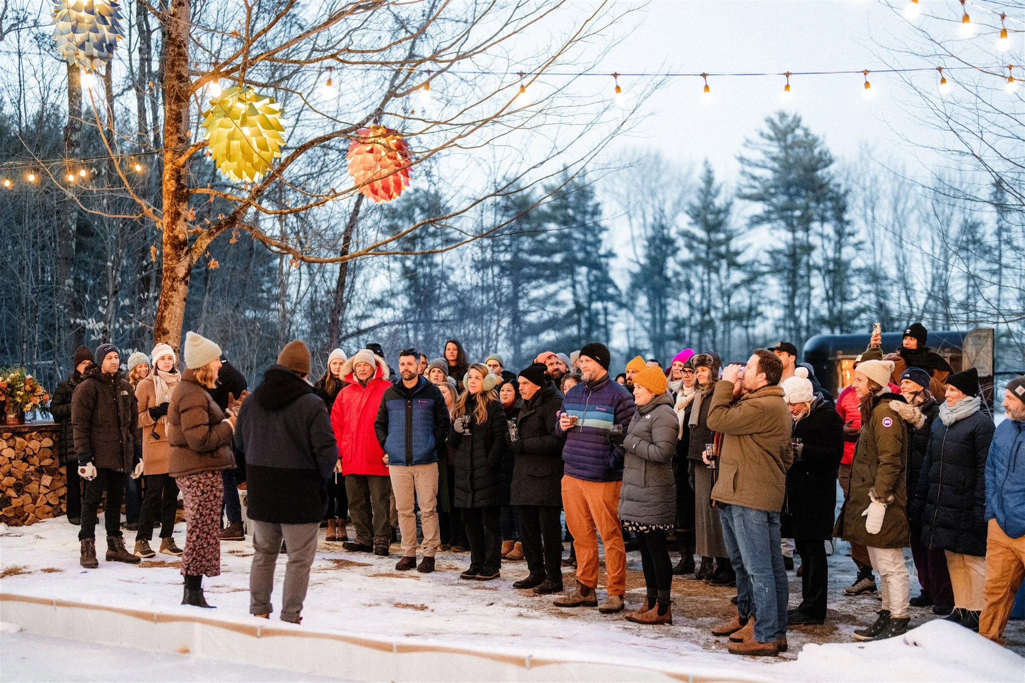 A group of people gathered outdoors in winter, wearing warm clothing with snow on the ground, under string lights and decorative paper lanterns hung from a tree.