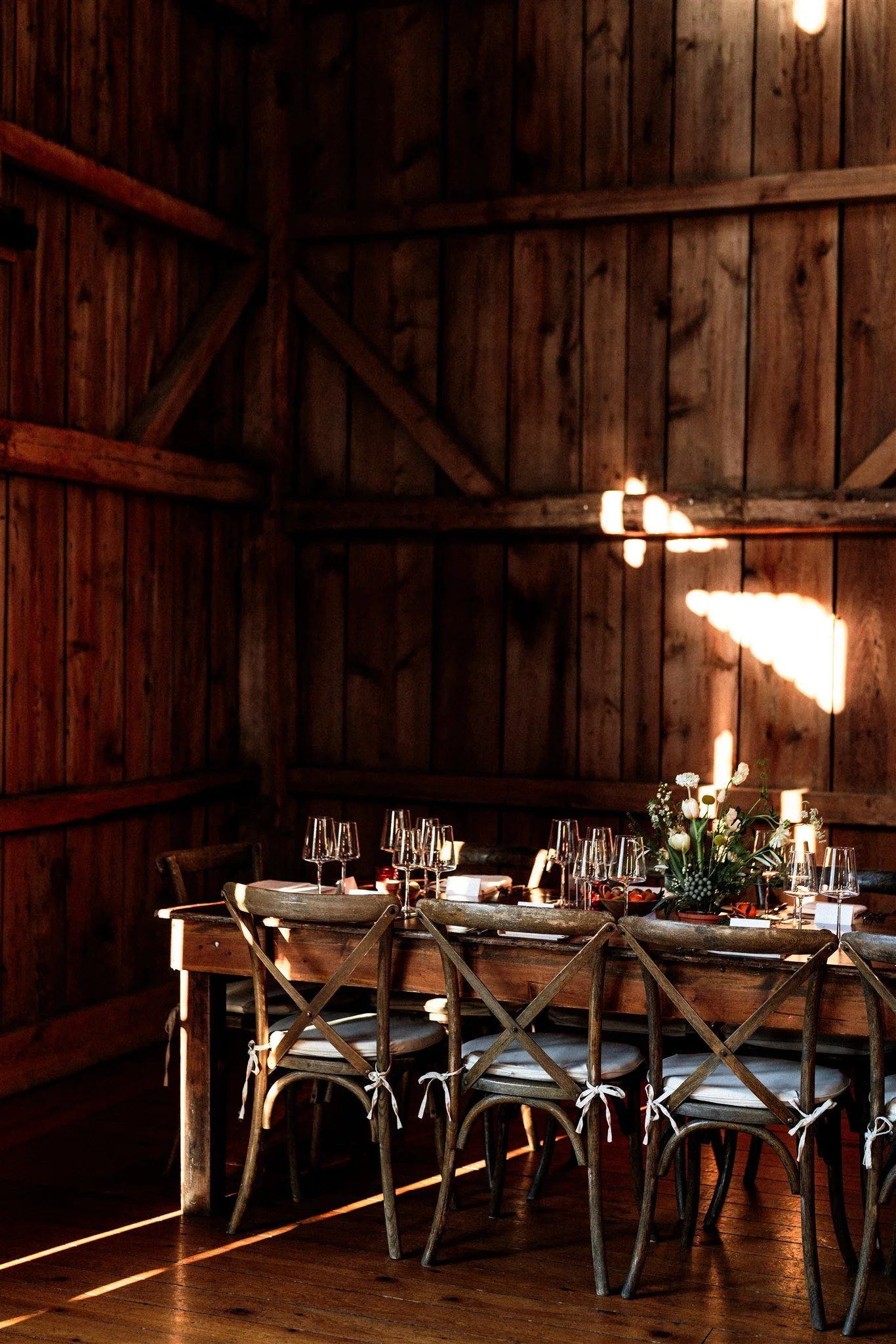 A rustic dining table set for a meal inside a wooden barn, with wine glasses, a flower centerpiece, and chairs with white cushions.