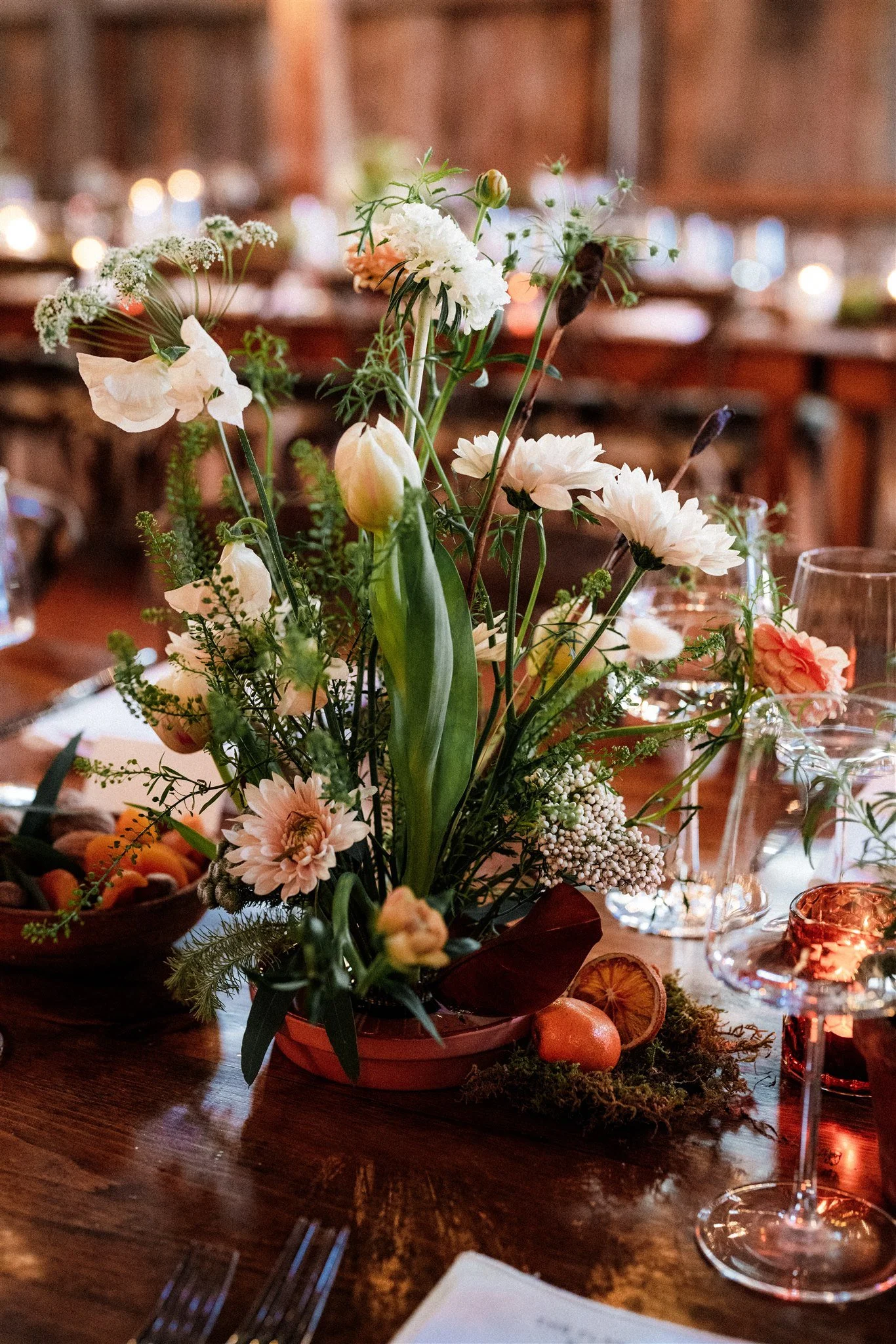 A floral centerpiece with white, pink, and green flowers on a wooden table, surrounded by glasses and tableware at an indoor event.