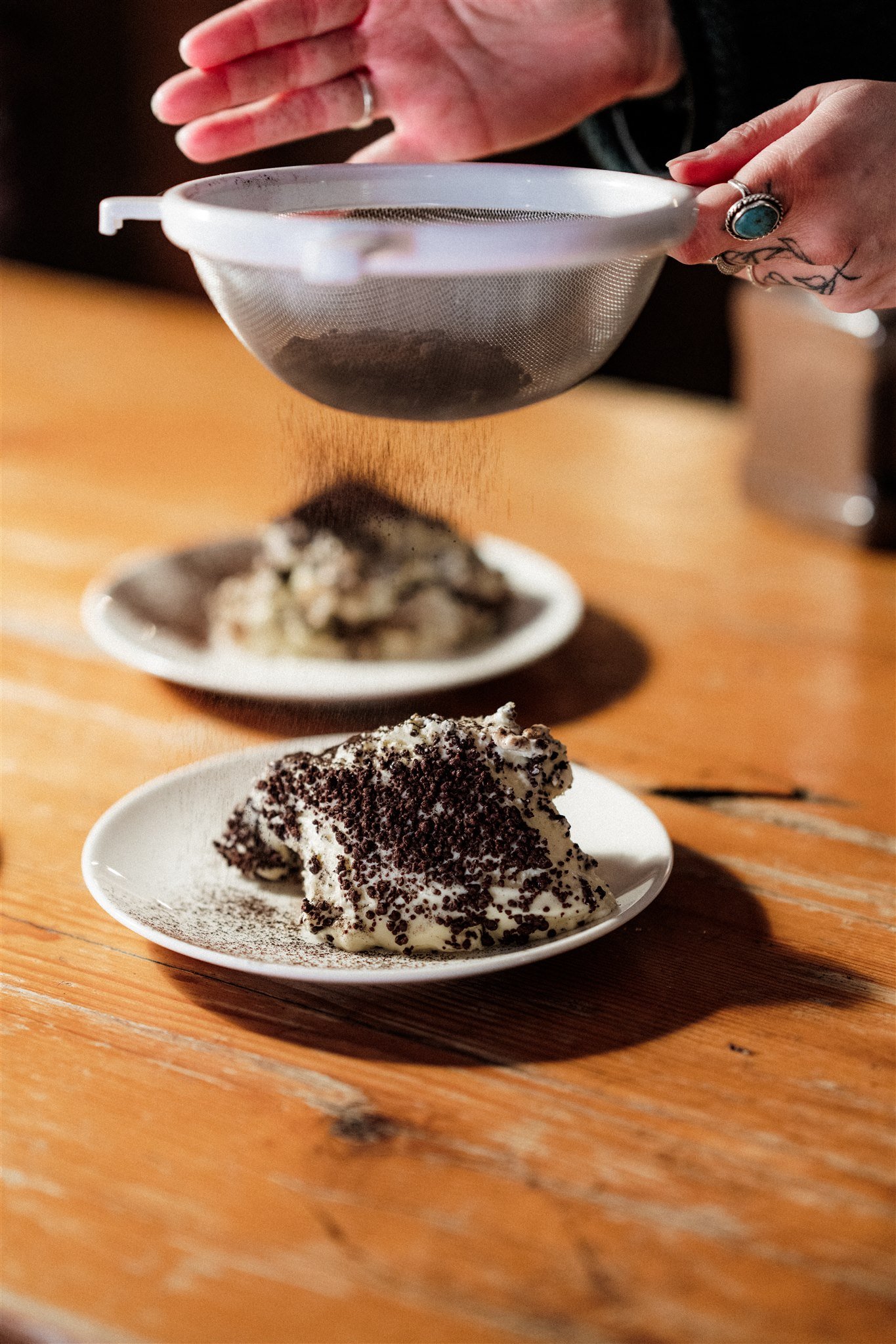 A person sifting cocoa powder over a bowl of ice cream topped with chocolate sprinkles on a wooden table.
