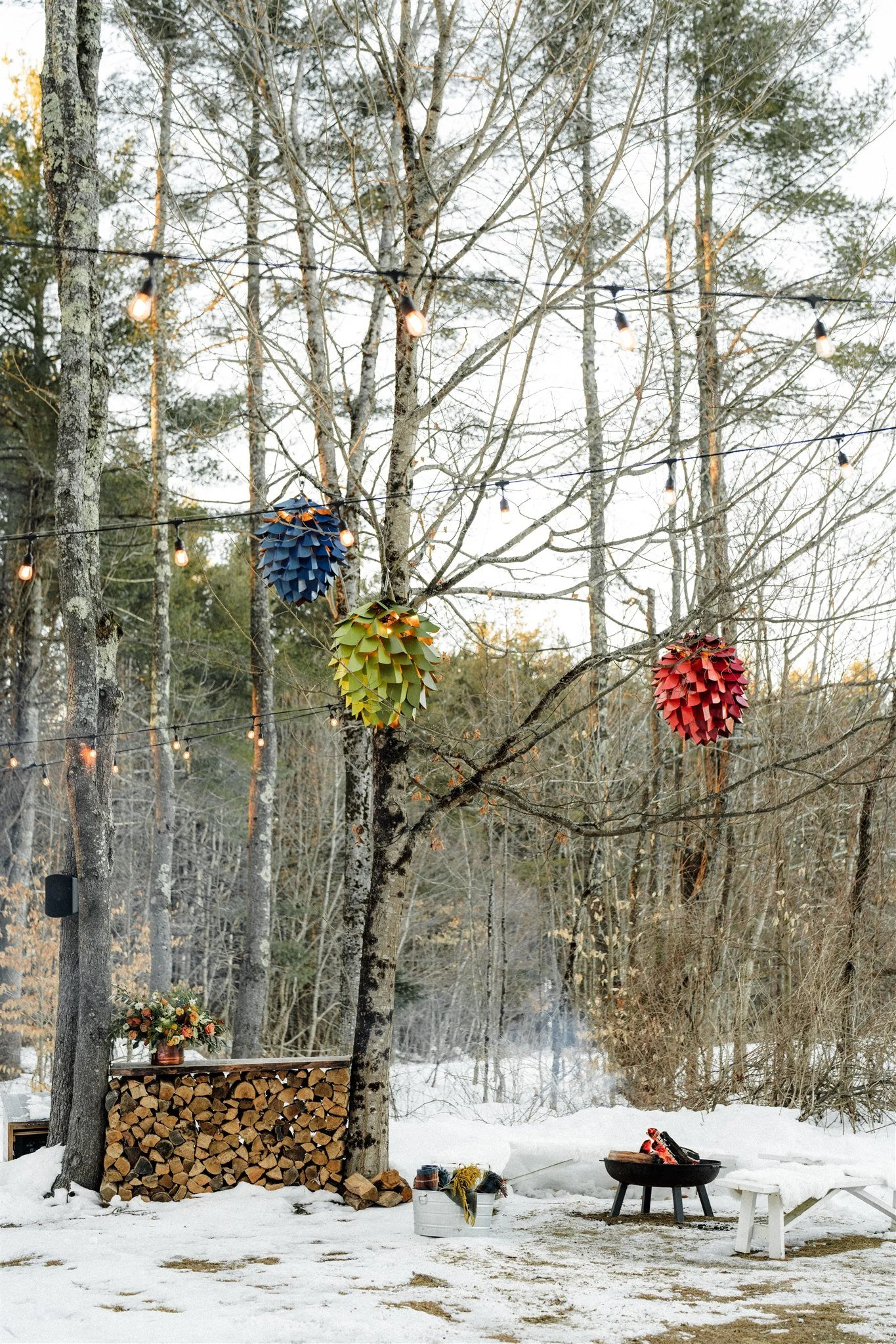 Outdoor winter scene with a bare tree decorated with hanging colorful paper lanterns and string lights, woodpile, flower arrangement, fire pit with logs, and small white chair.