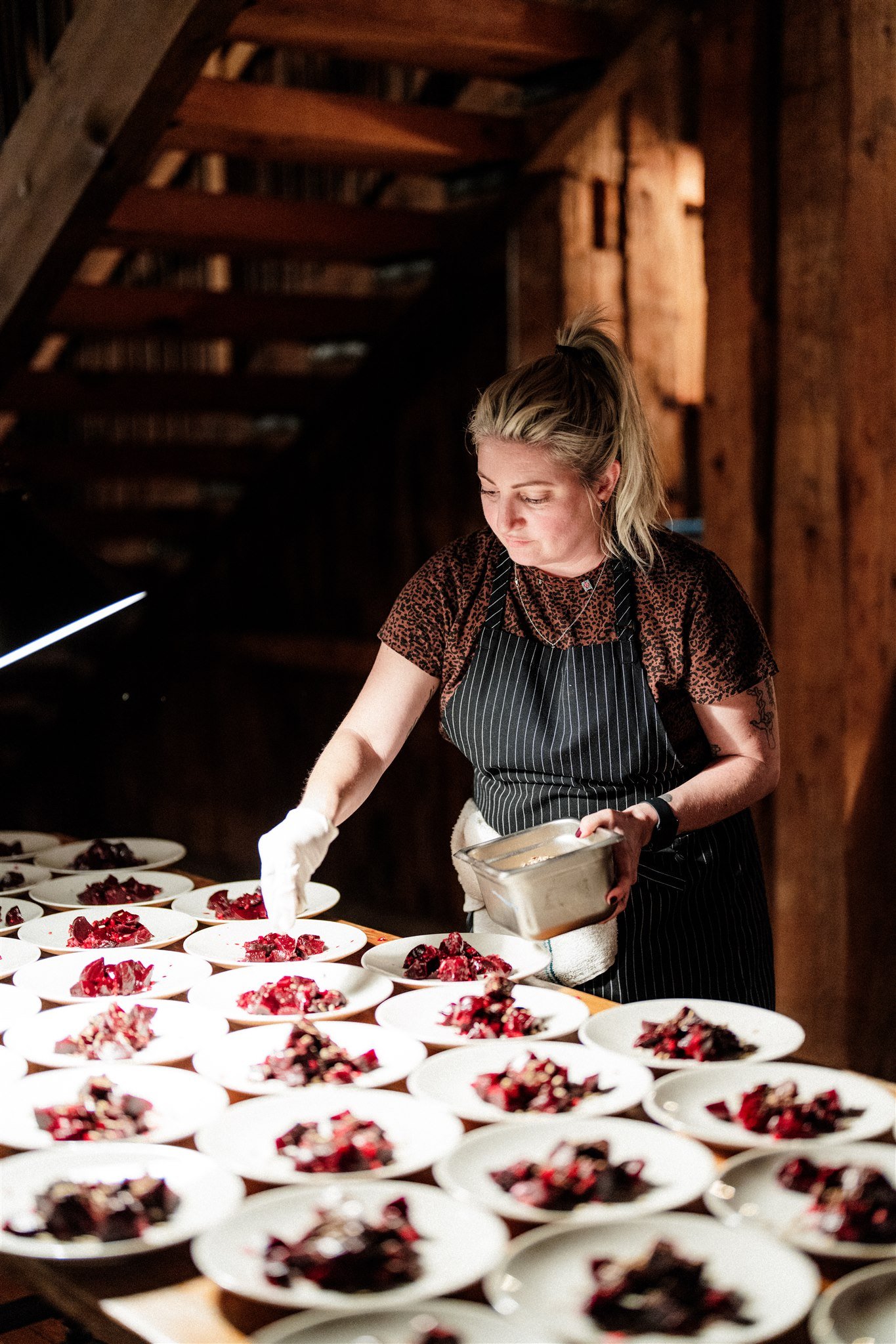 A woman in a black apron and white glove preparing multiple plates of a red dessert in a rustic, wood-paneled kitchen or dining area.