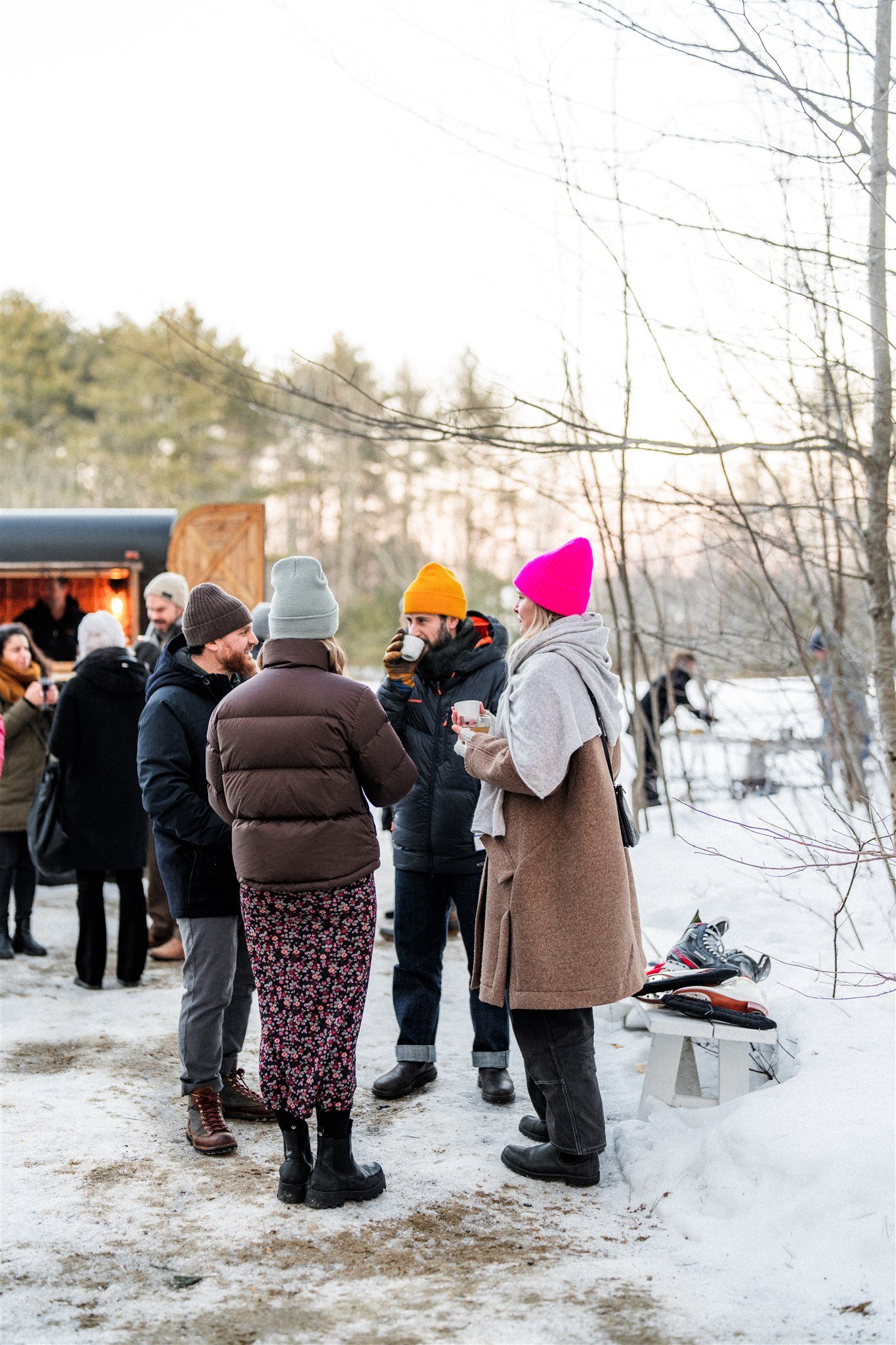 Group of people socializing outdoors in a snowy setting, some holding drinks, with a food truck in the background.