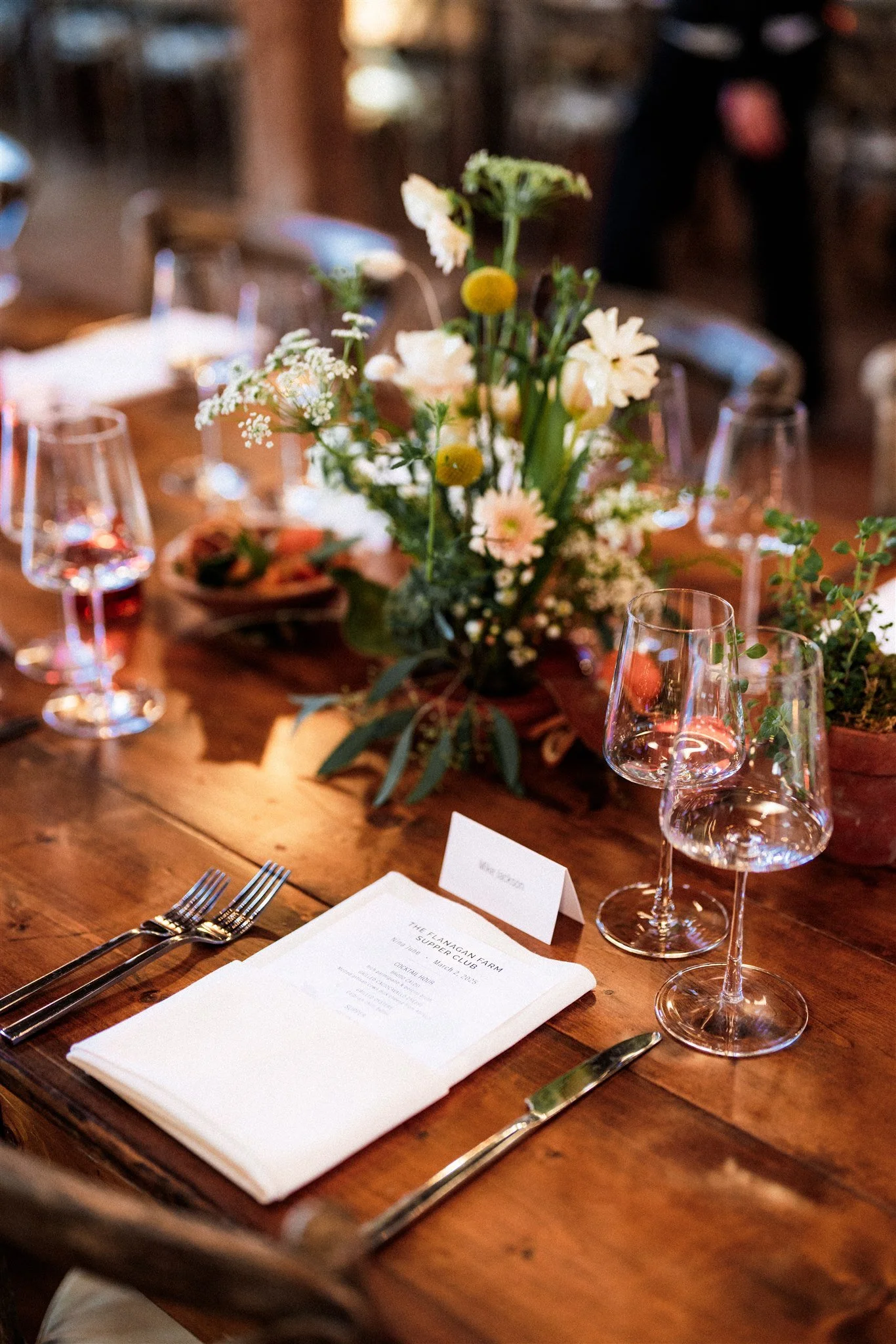 A rustic dinner table setting with a floral centerpiece, wine glasses, and a white folded napkin with printed text, in a warm, wooden interior.