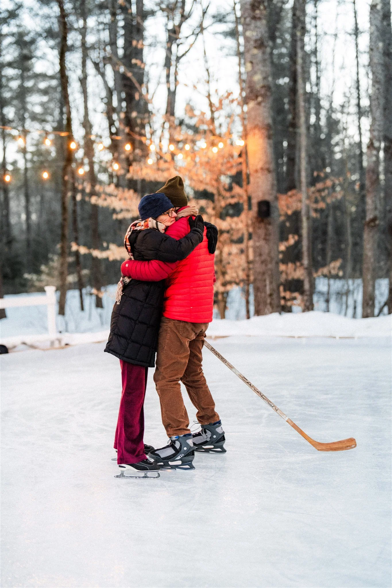 Two people in winter clothing hugging on an outdoor ice skating rink with trees and string lights in the background.