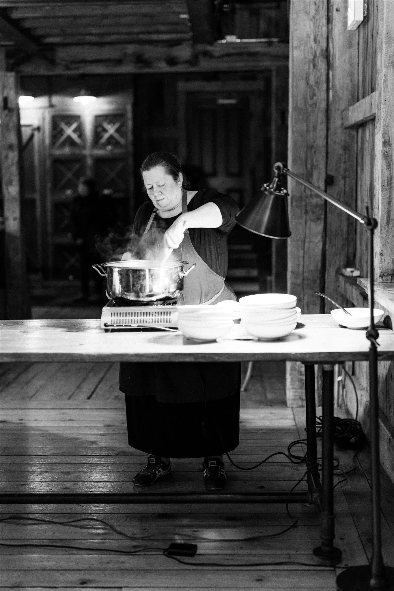 A woman cooking on a stove in a rustic setting, with stacked bowls on the table and a lamp illuminating her workspace.