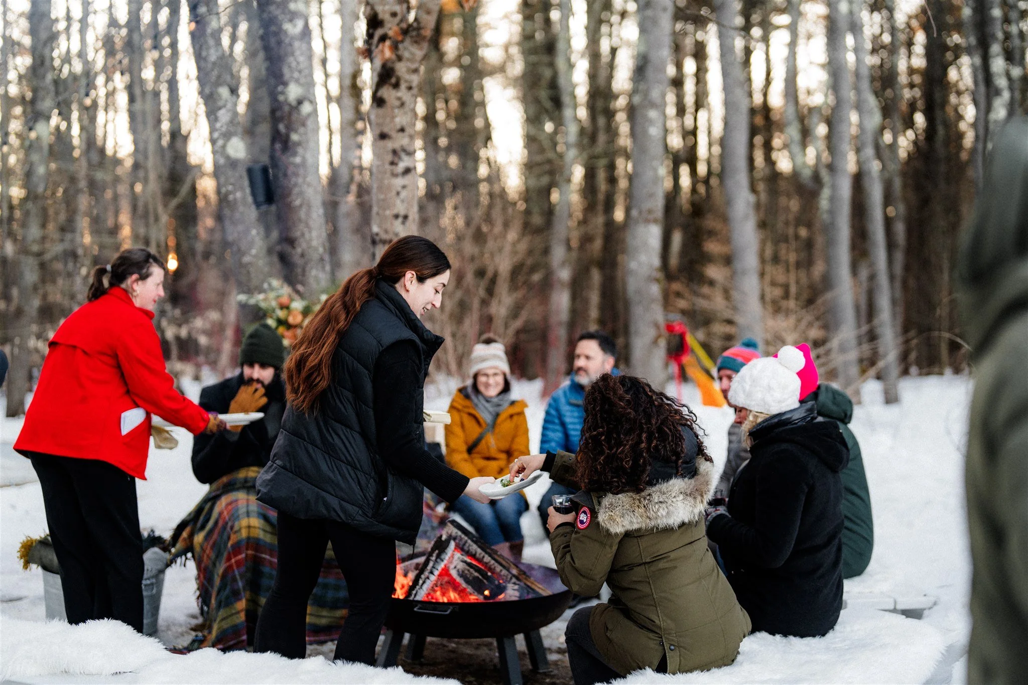 Group of people gathered around a campfire in a snowy forest, enjoying food and drinks outdoors in winter.