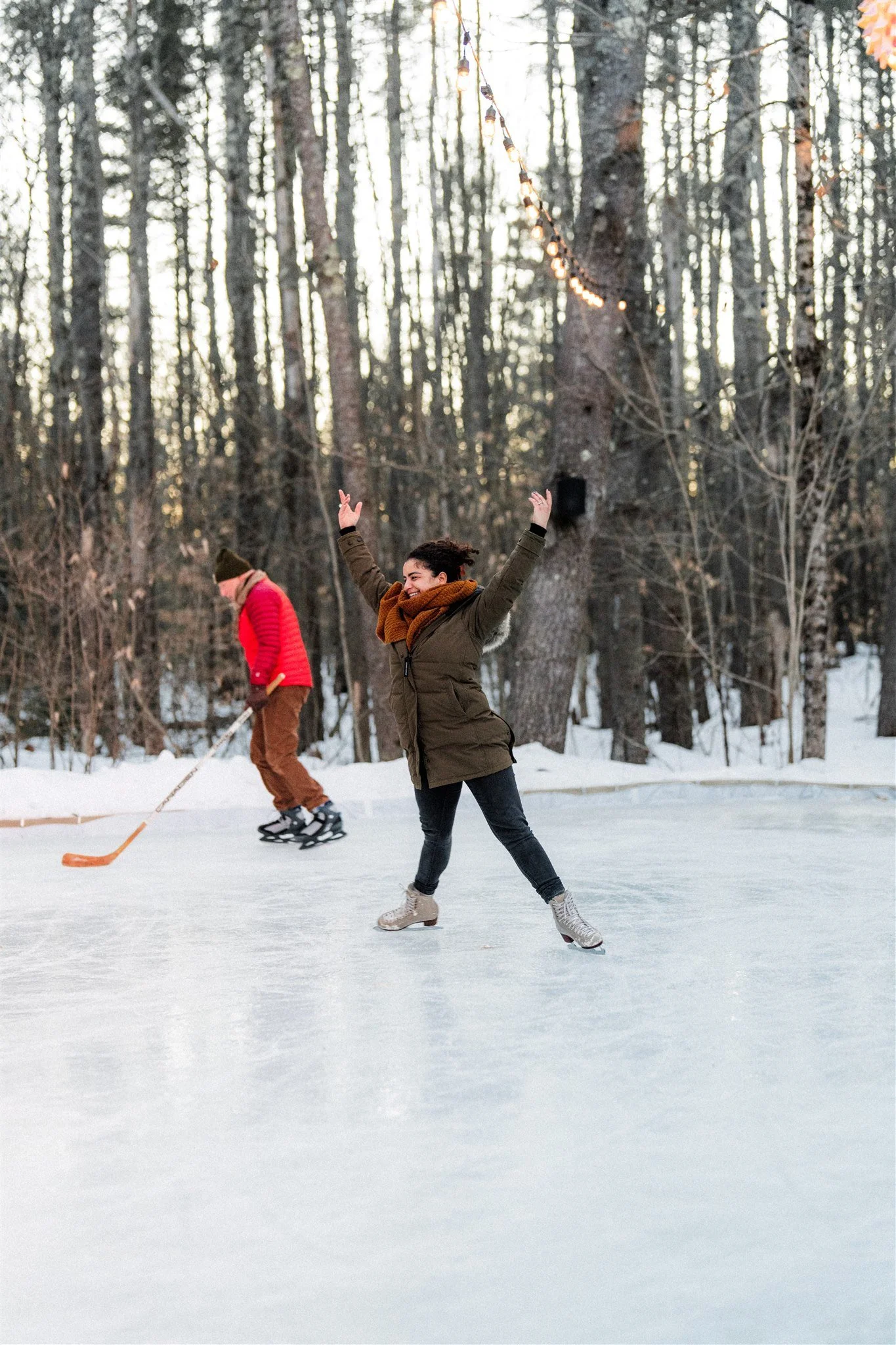 Two people ice skating on an outdoor rink in a winter forest during sunset, one smiling with arms raised, the other in the background using a hockey stick.