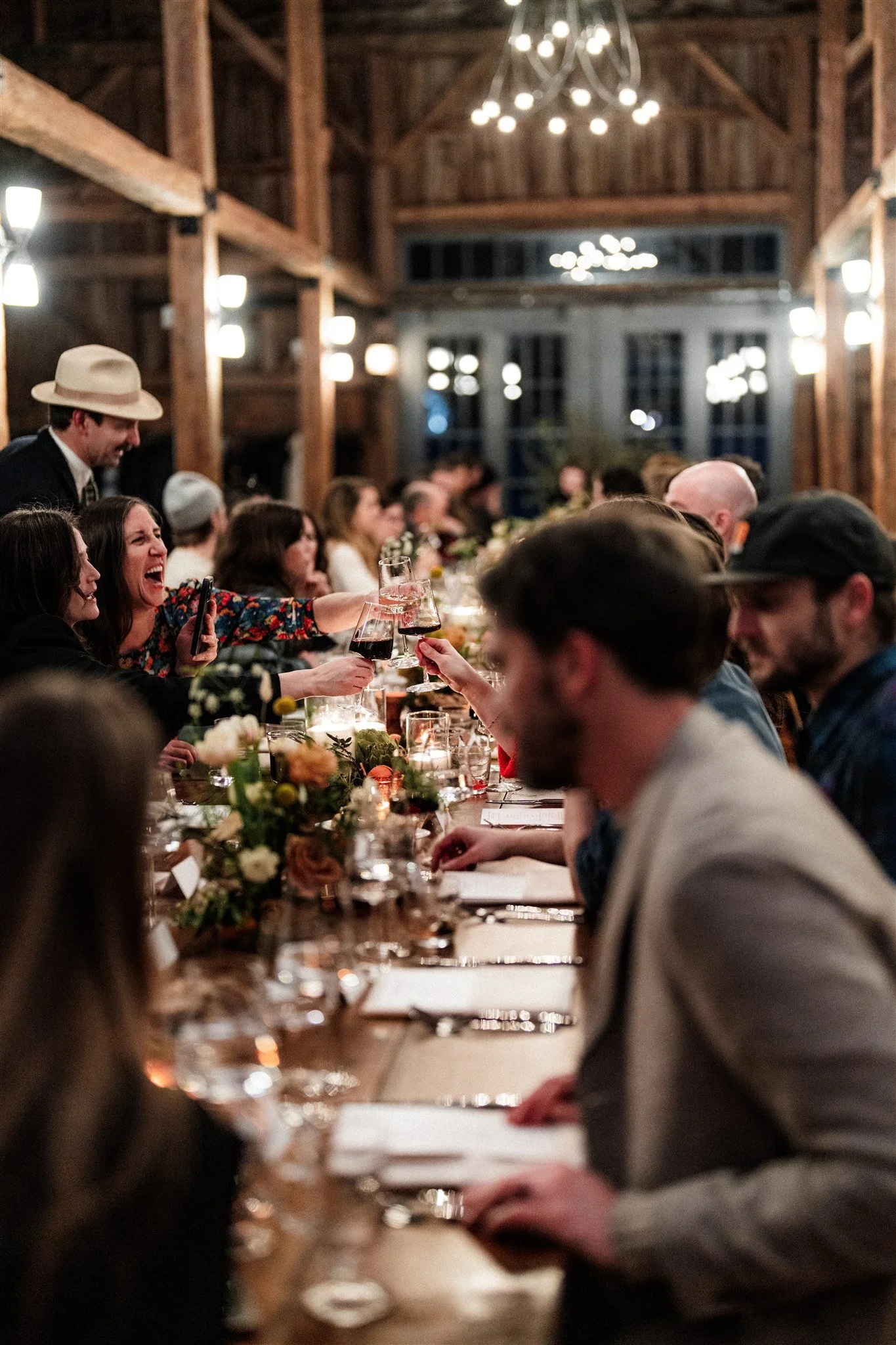 People sitting at a long dinner table in a rustic wooden venue, raising glasses in a toast, with floral centerpieces and candles.