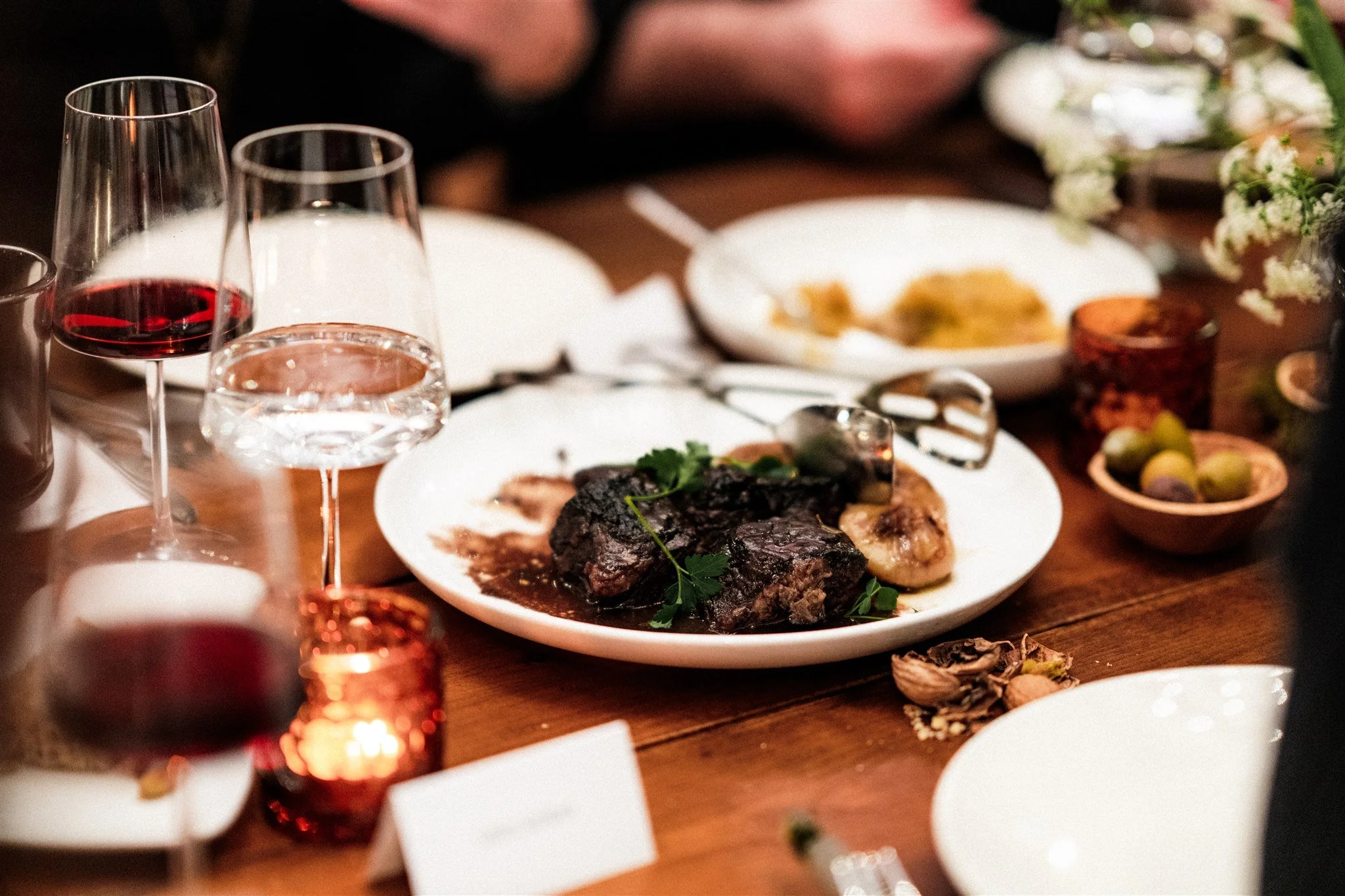 A dinner table with glasses of red and white wine, and plates of food, including a main dish of dark braised meat with herbs, and side dishes with vegetables and bread, decorated with flowers and candles.