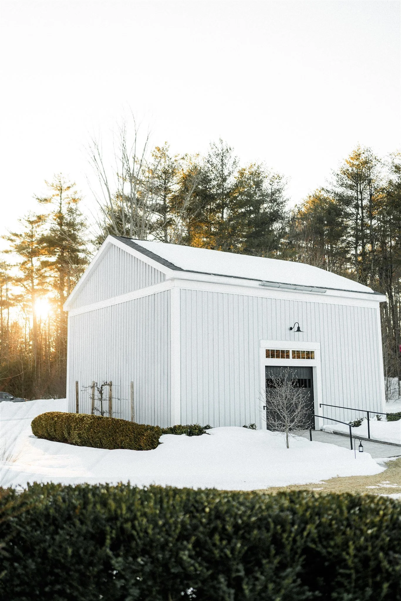 A white barn with black accents in a snowy landscape, with trees and the setting sun in the background.