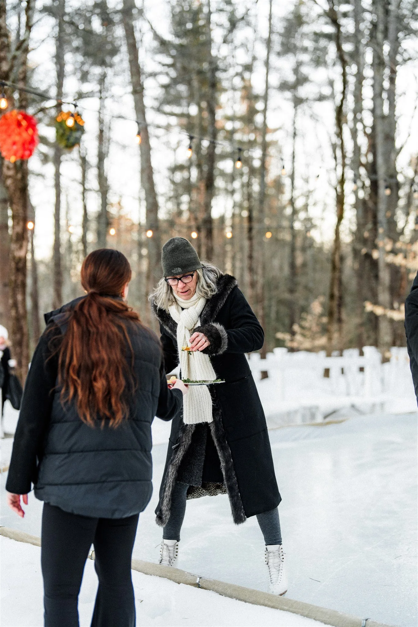 Two women talking outdoors in winter, with one holding a tray of food, surrounded by snow and trees under string lights.