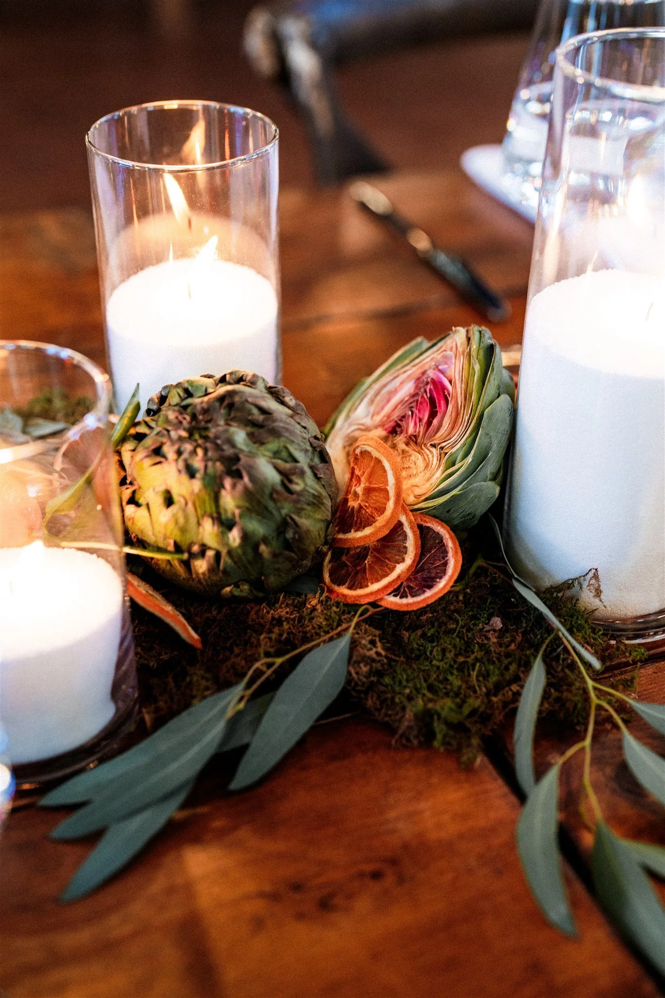 Decorative table centerpiece with candles, artichoke, dried orange slices, and greenery on a wooden table.