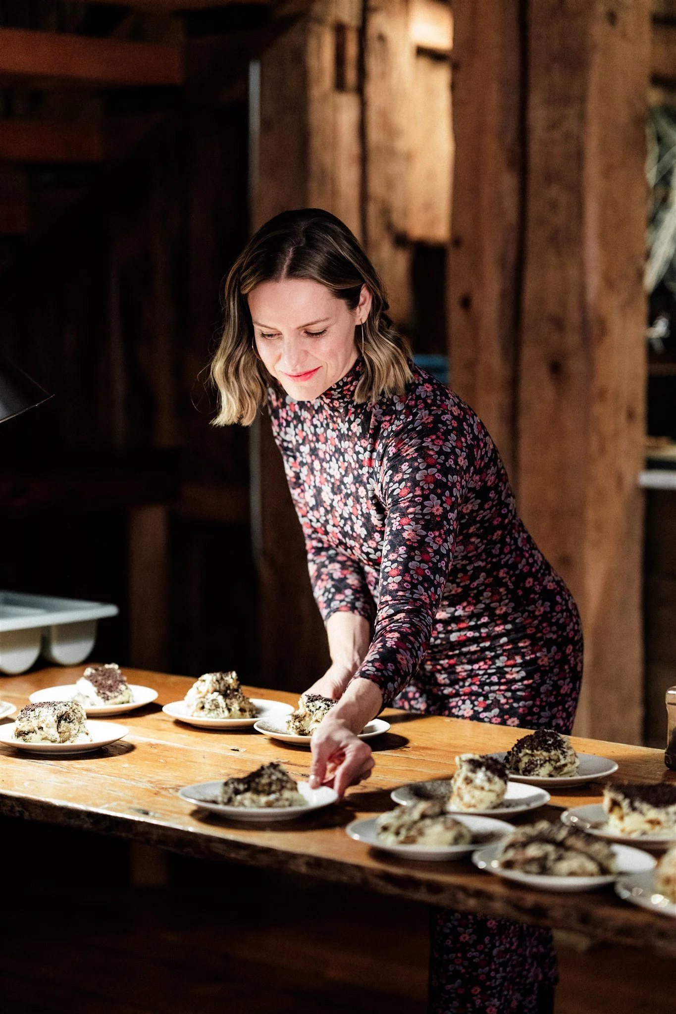 A woman arranging plates of cake on a wooden table in a cozy, rustic setting.