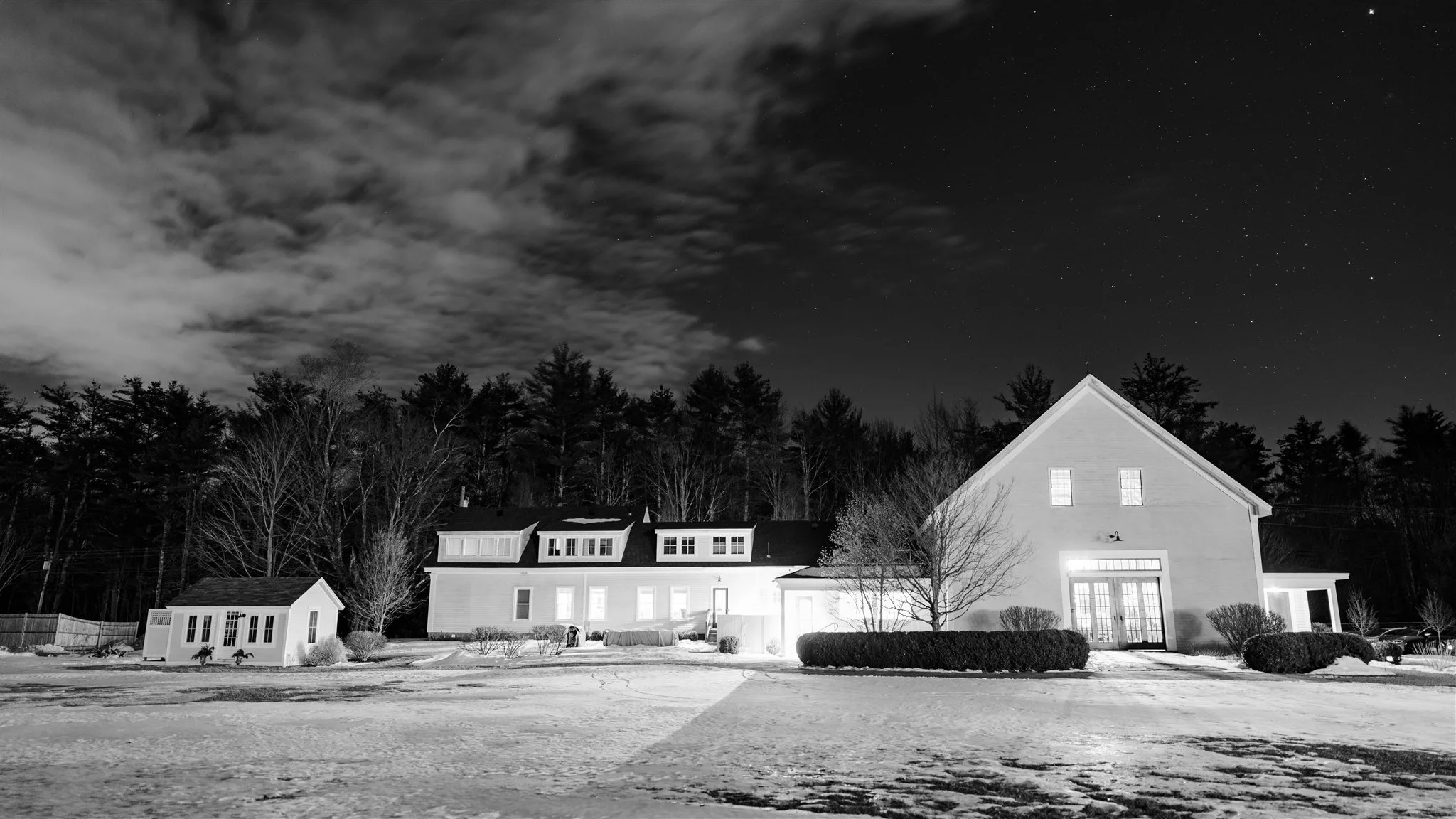 A large house with illuminated windows and a smaller building in front, both surrounded by trees and snow-covered ground at night, under a cloudy sky with stars.