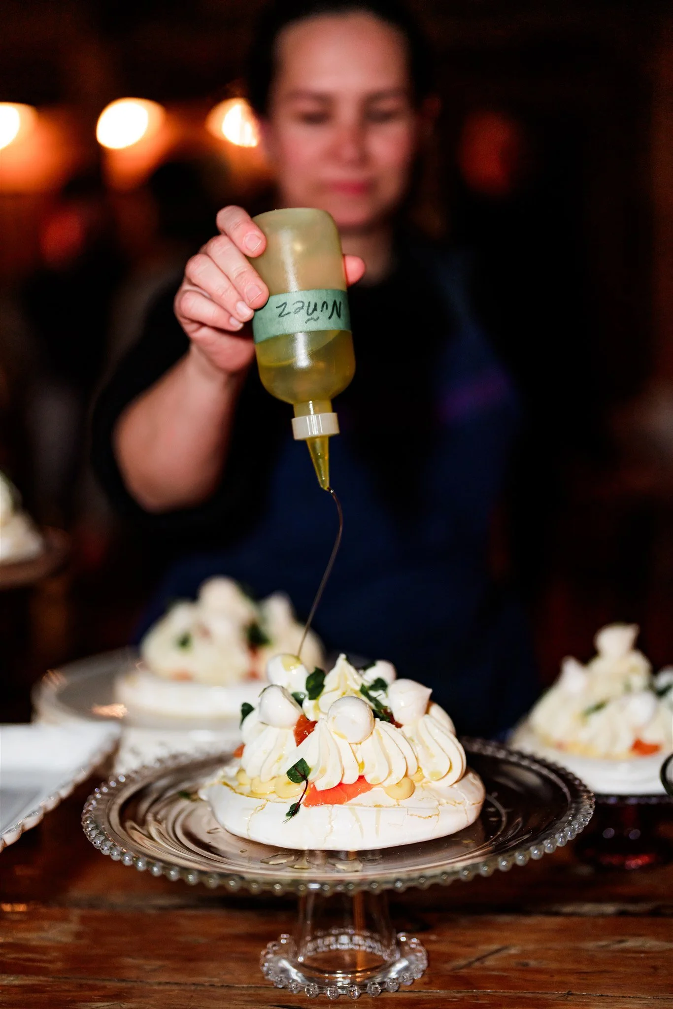 A person is holding a squeeze bottle of yellow sauce, pouring it onto a pavlova dessert topped with whipped cream, berries, and garnishes.