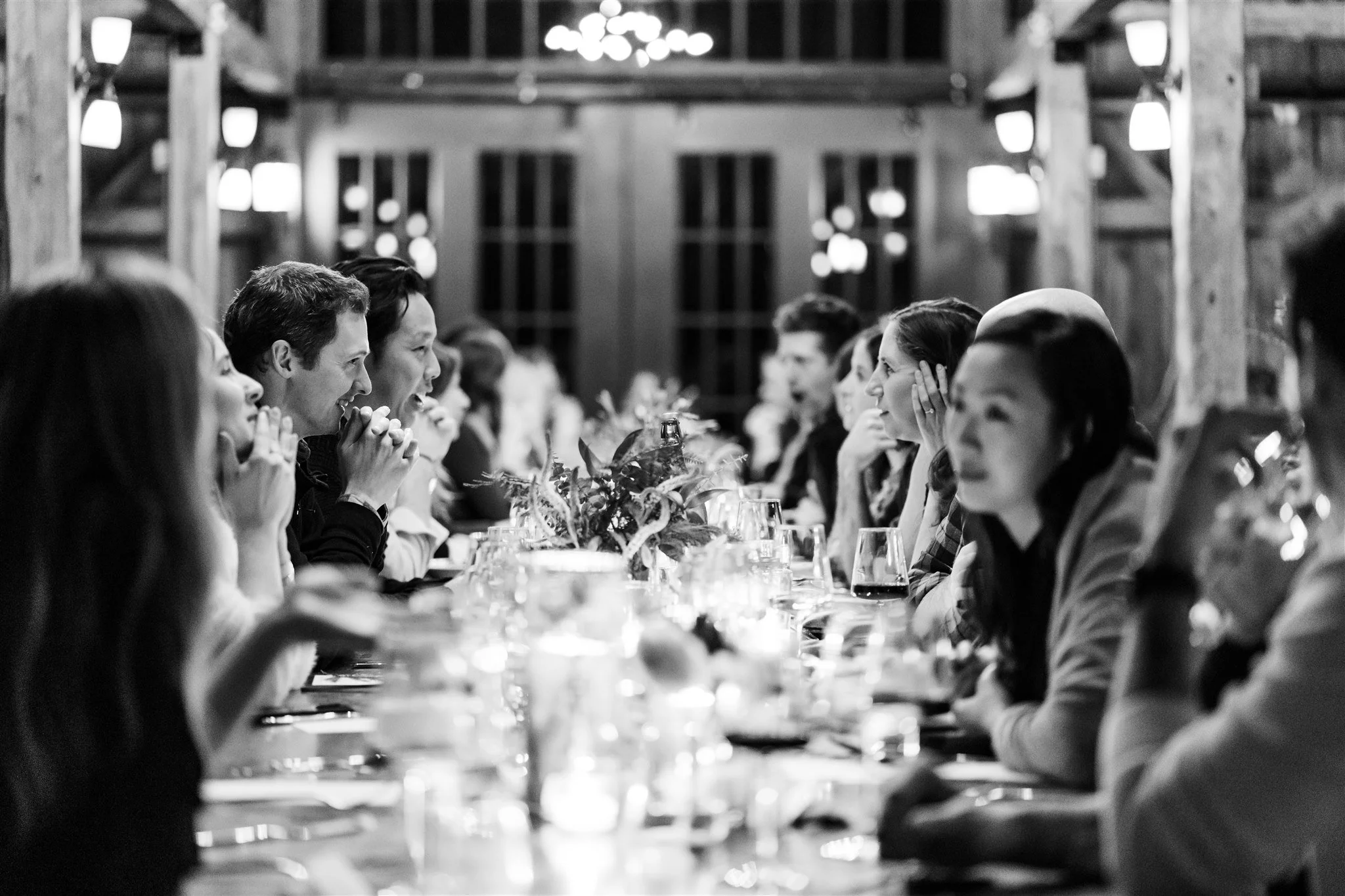 People sitting at a long dining table in a rustic barn, enjoying a meal and conversation, decorated with flowers and wine glasses.