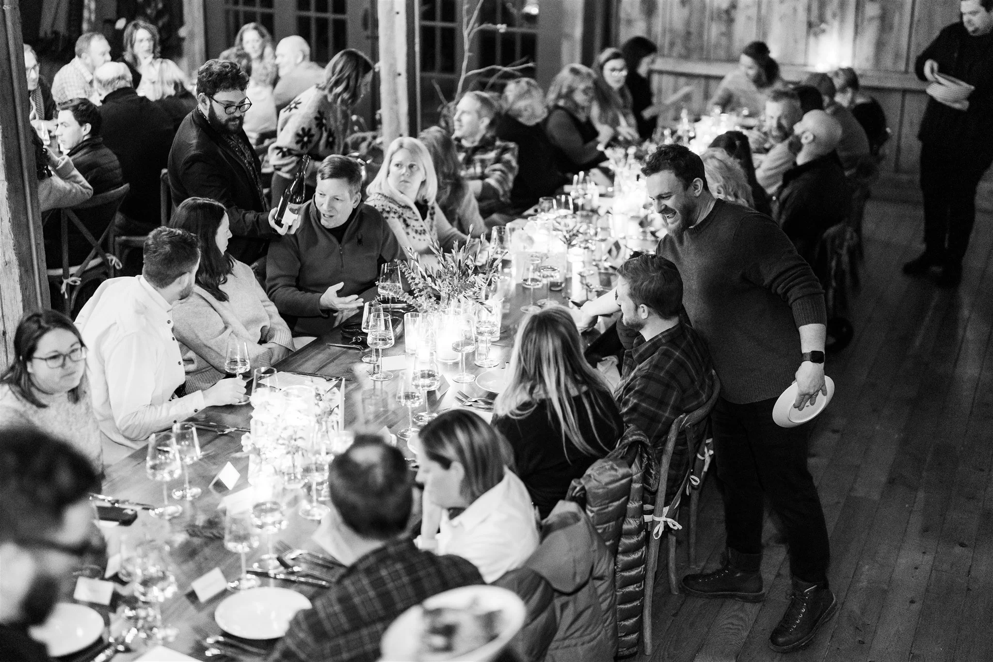 People gathered around a long dining table at a social event, engaging in conversation and enjoying drinks, in a rustic indoor setting.