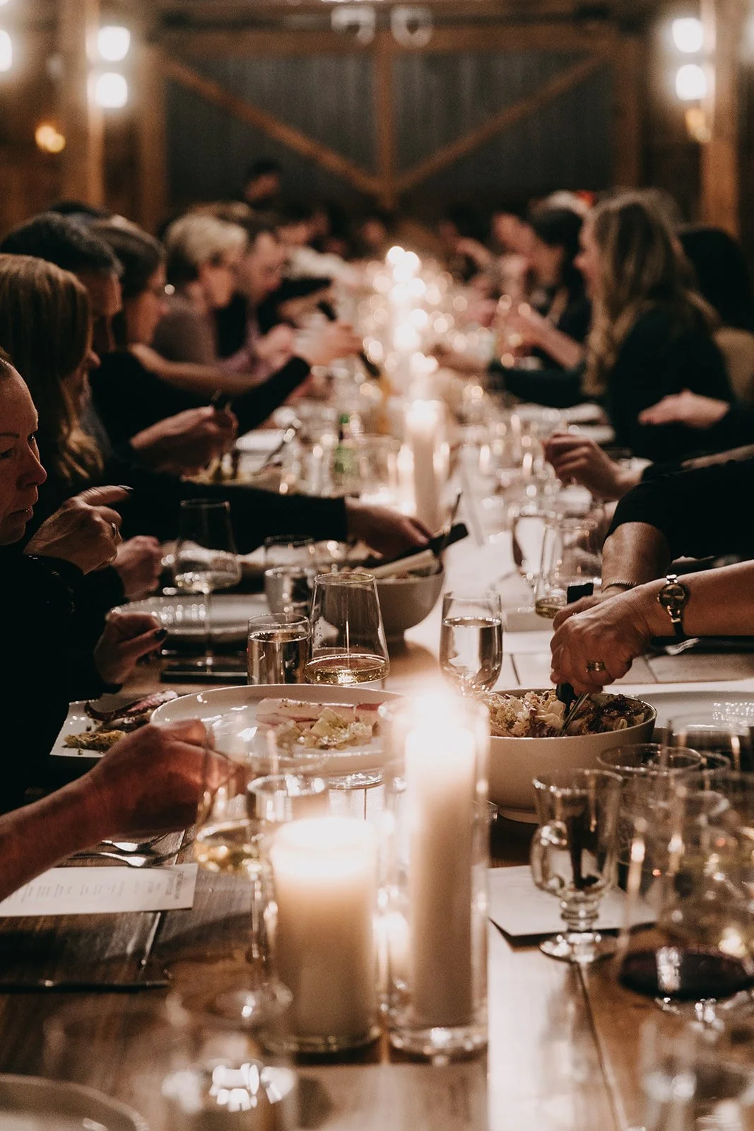 A long dinner table crowded with people eating, drinking, and socializing in a rustic, warmly lit setting with candles.