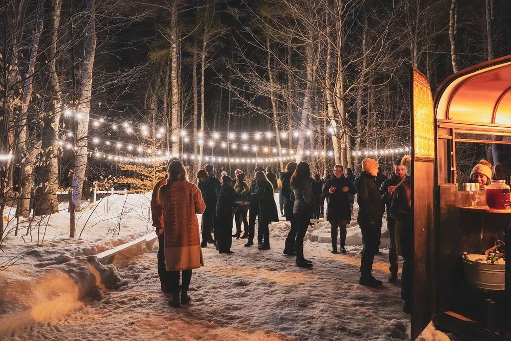 People gathered outdoors at night in a snowy forest, illuminated by string lights, with a food or drink trailer nearby.