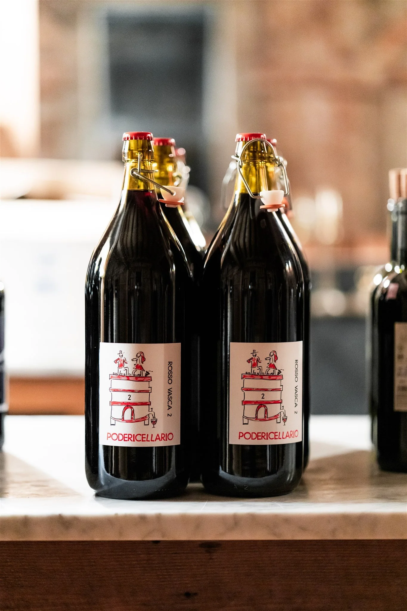 Four bottles of red wine with swing-top caps on a wooden surface, labels feature a drawing of a tower with two figures, text includes 'Rosso Vasca 2' and 'Podericellario'.