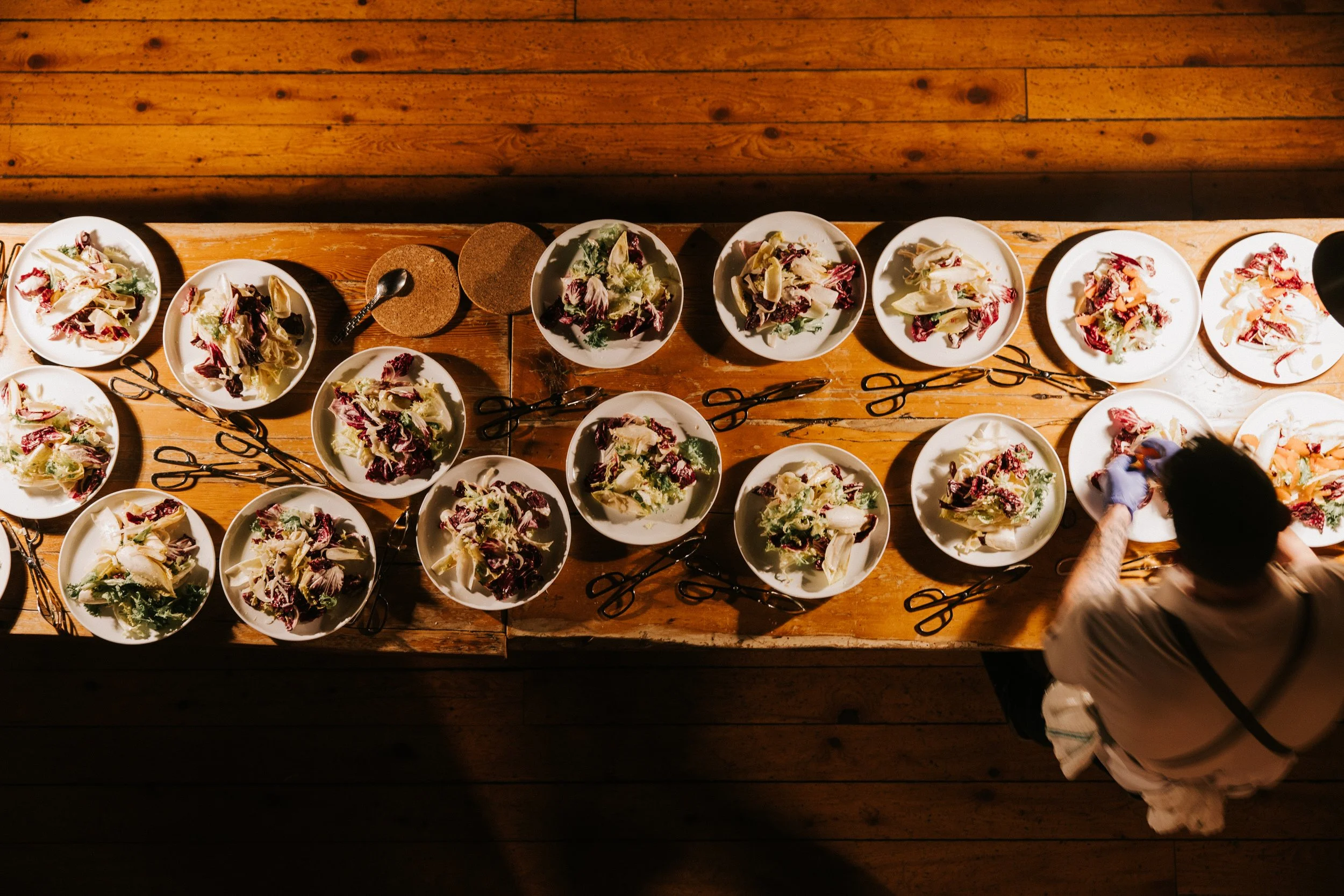 A person preparing salads on a wooden table with multiple plates of mixed greens and utensils.