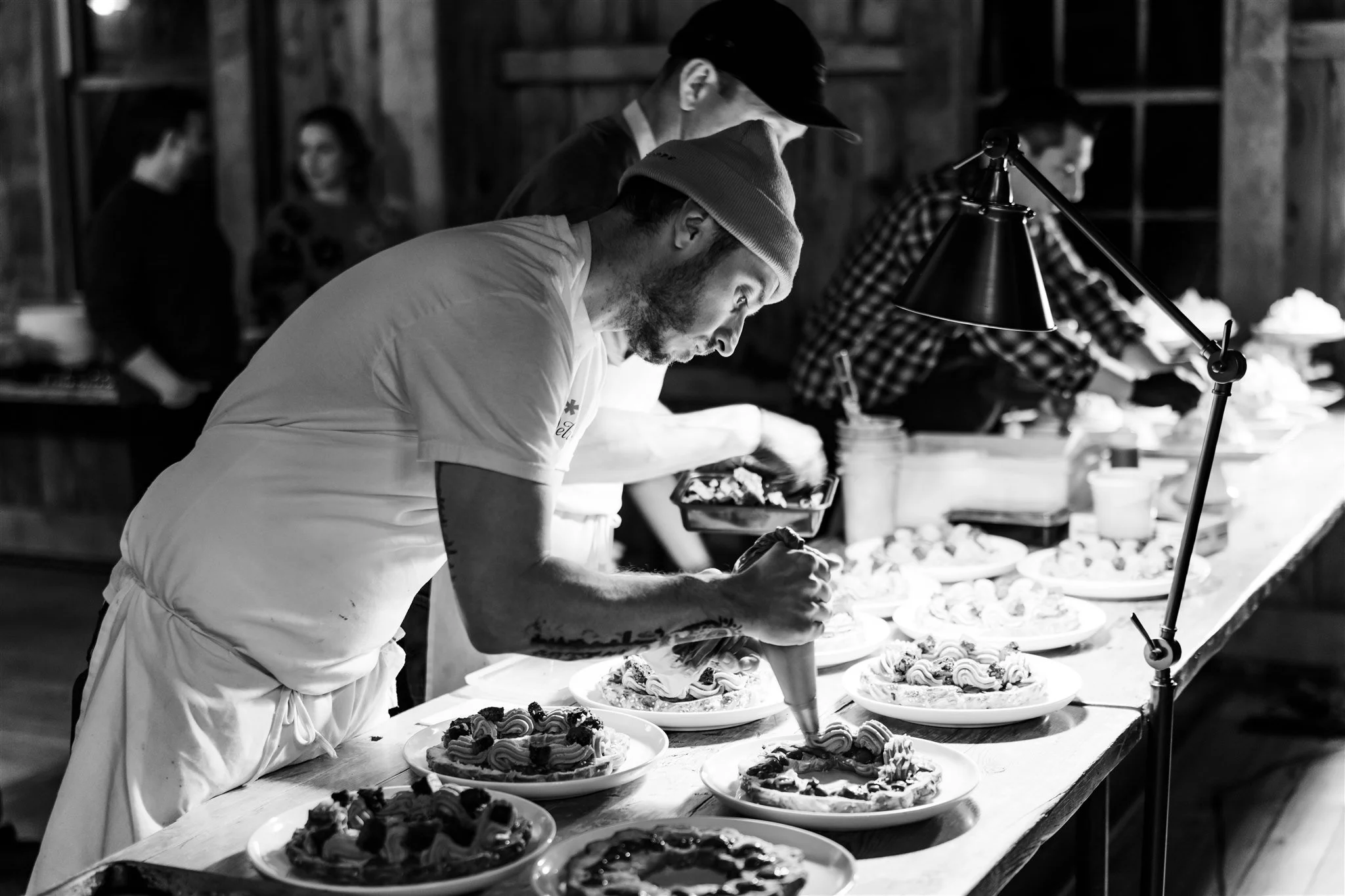 A chef decorates a fruit tart with whipped cream at a bakery counter with multiple tarts displayed, in a rustic wooden interior, in black and white.