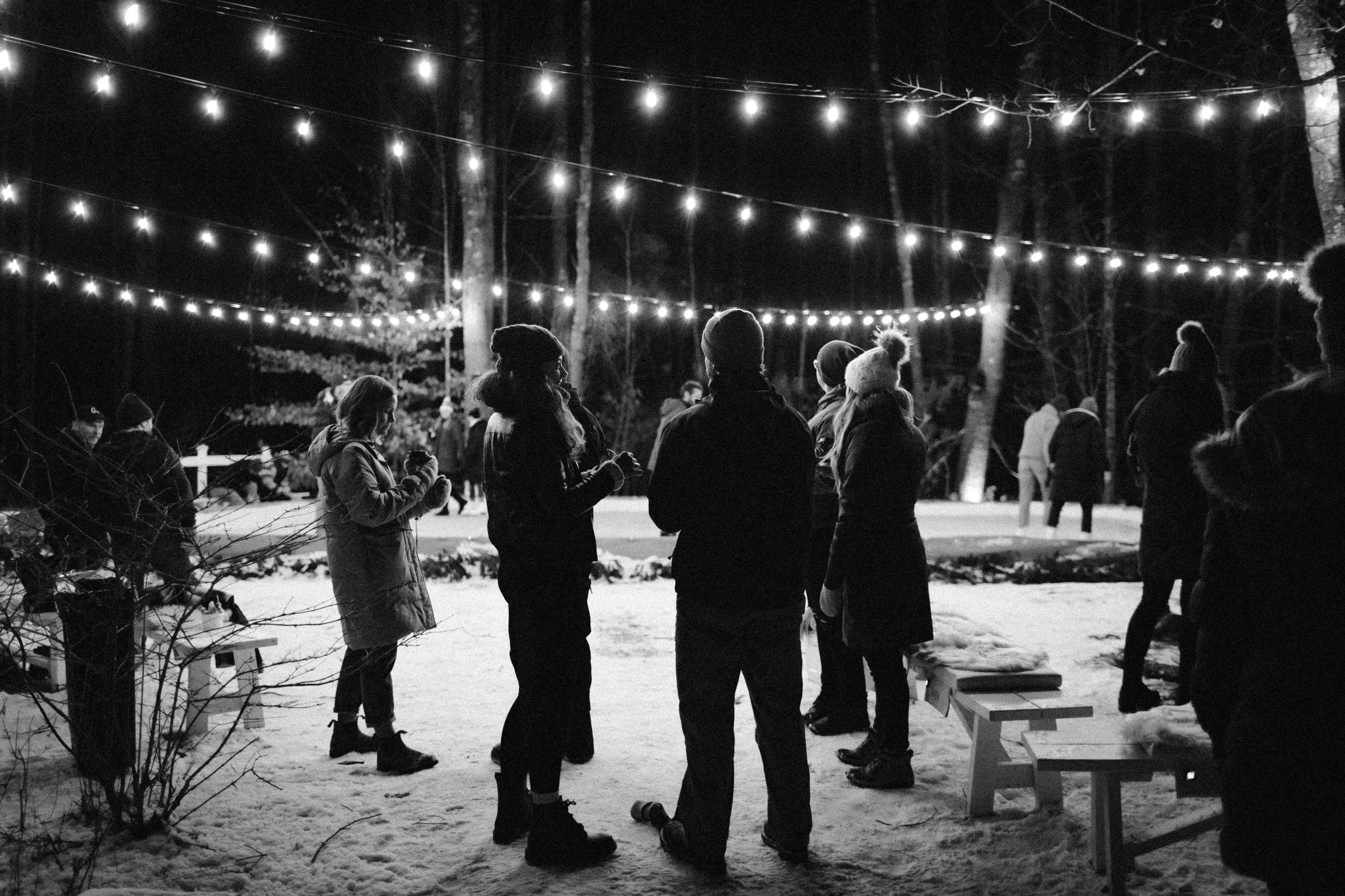 People gathering outdoors at night, illuminated by string lights, in a snowy wooded area.