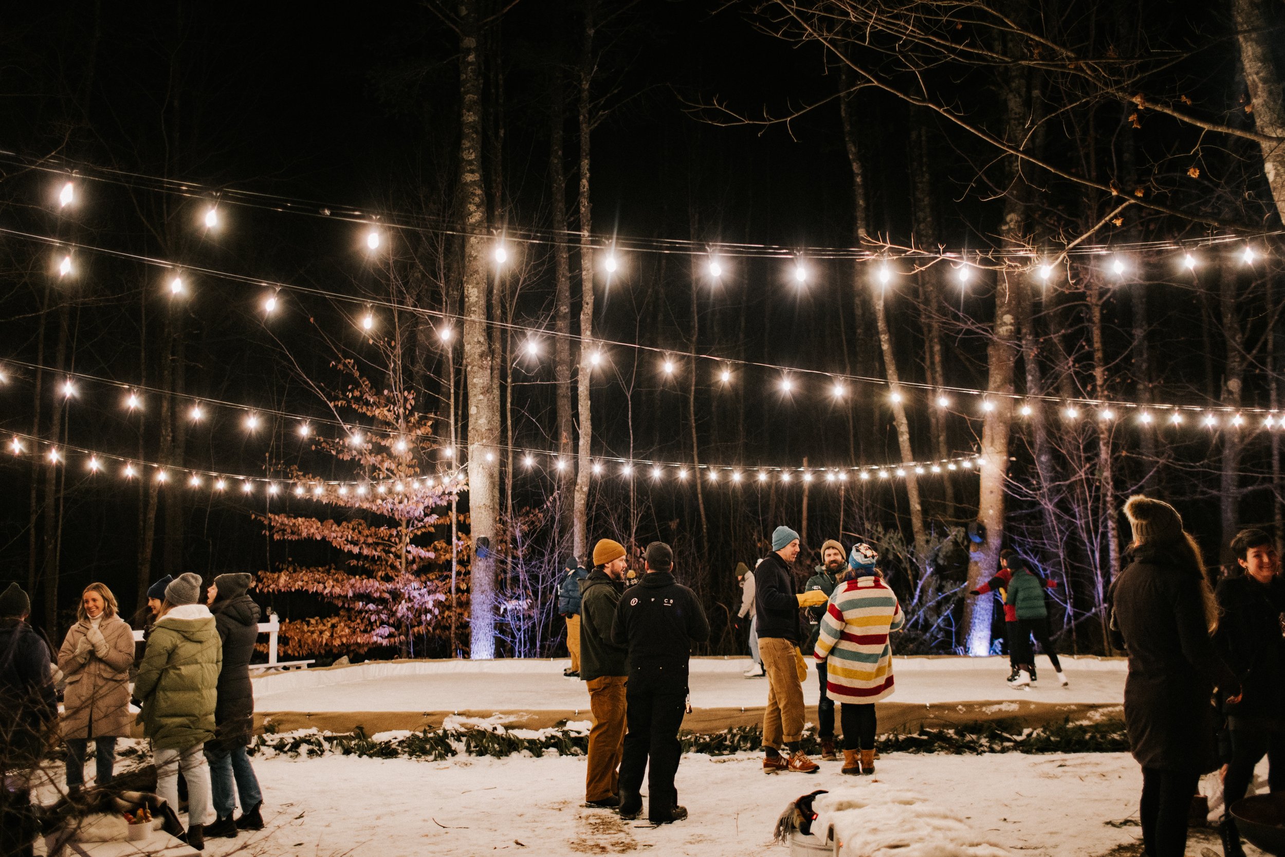 People enjoying an outdoor ice skating rink under string lights at night in a forested area, with snow on the ground and trees in the background.