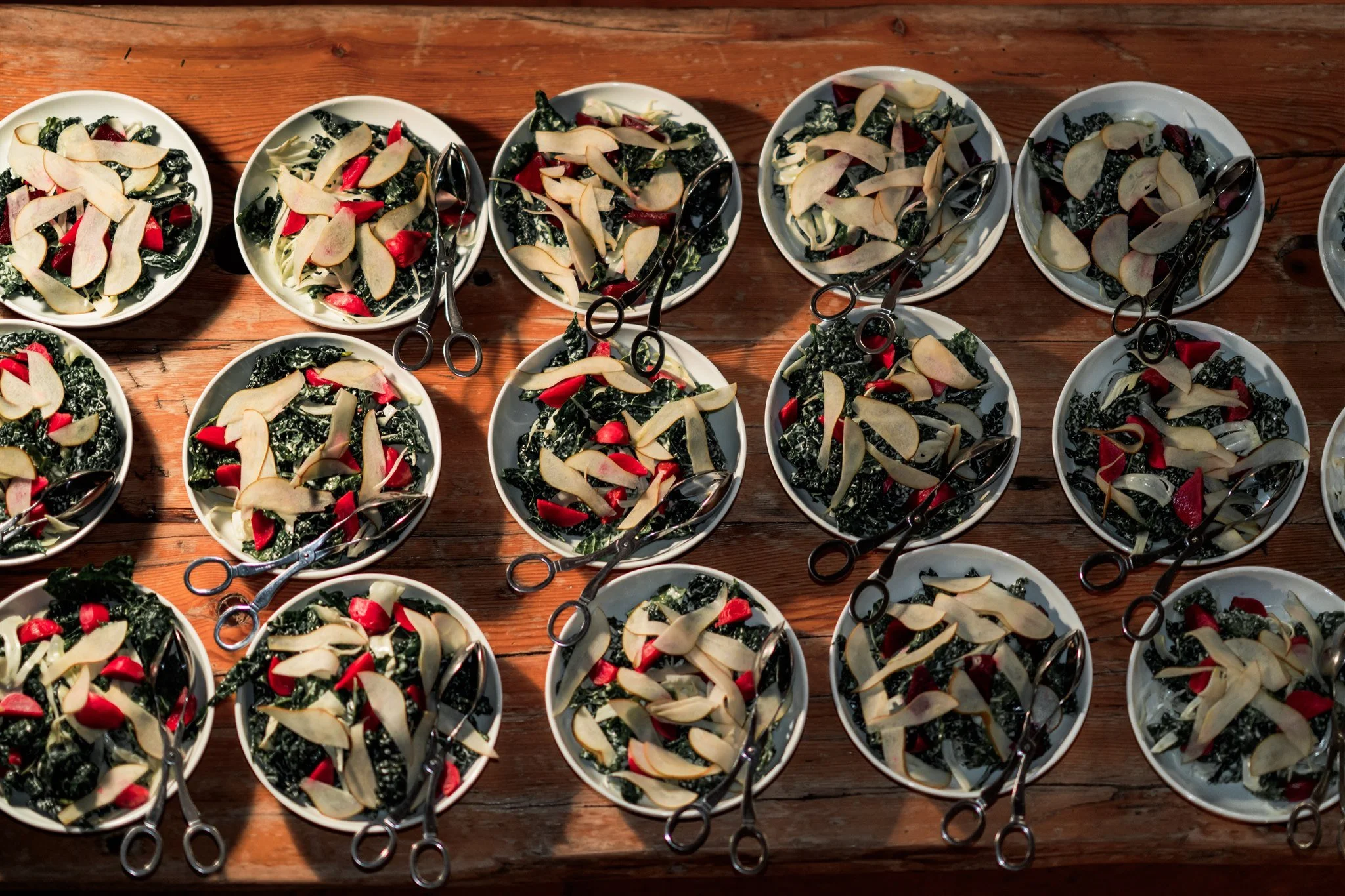 Multiple bowls of salad with kale, radishes, and sliced apples on a wooden table, each with a pair of tongs.