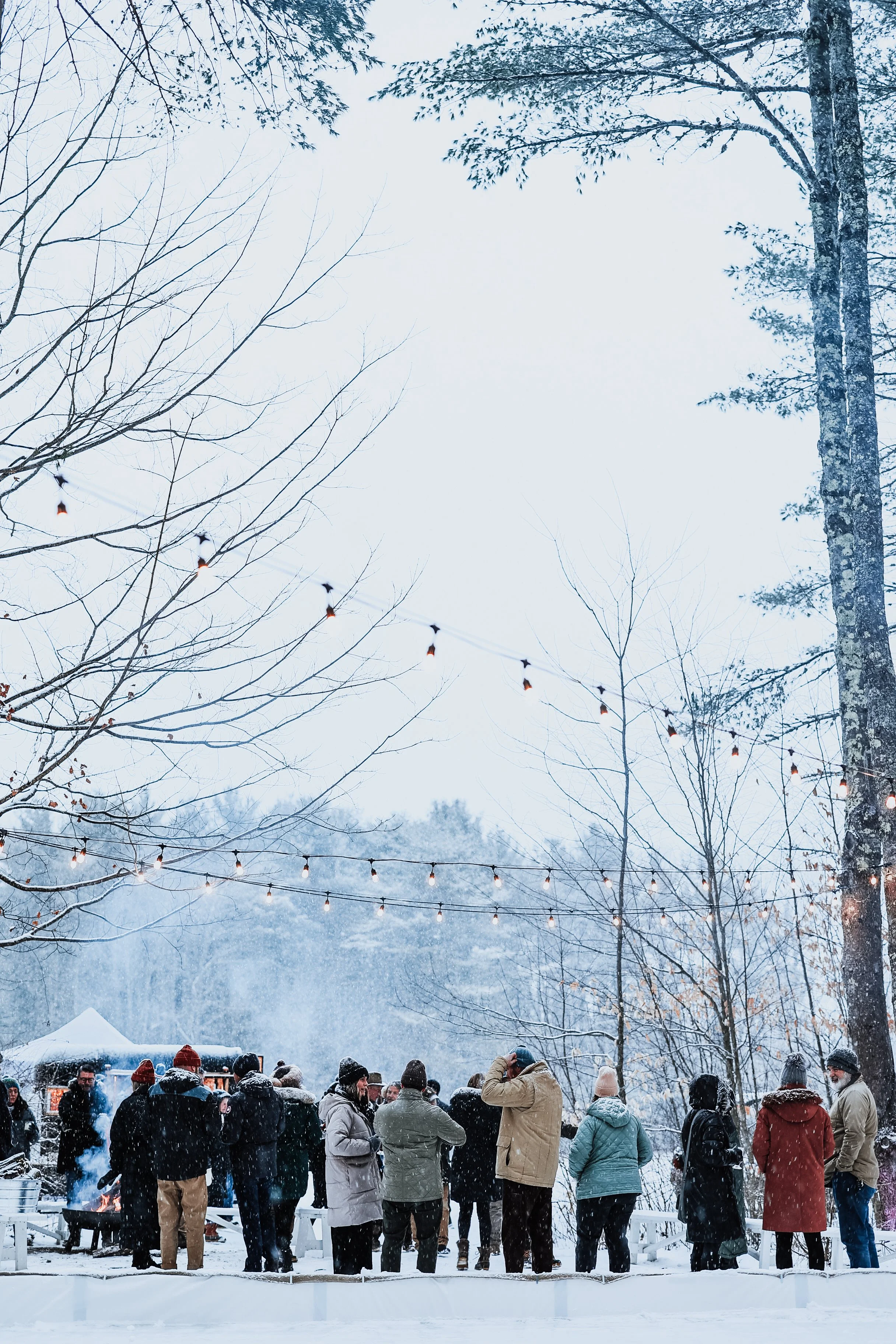A group of people gathered outdoors in snowy winter weather, under string lights, with trees and a small tent or booth in the background.