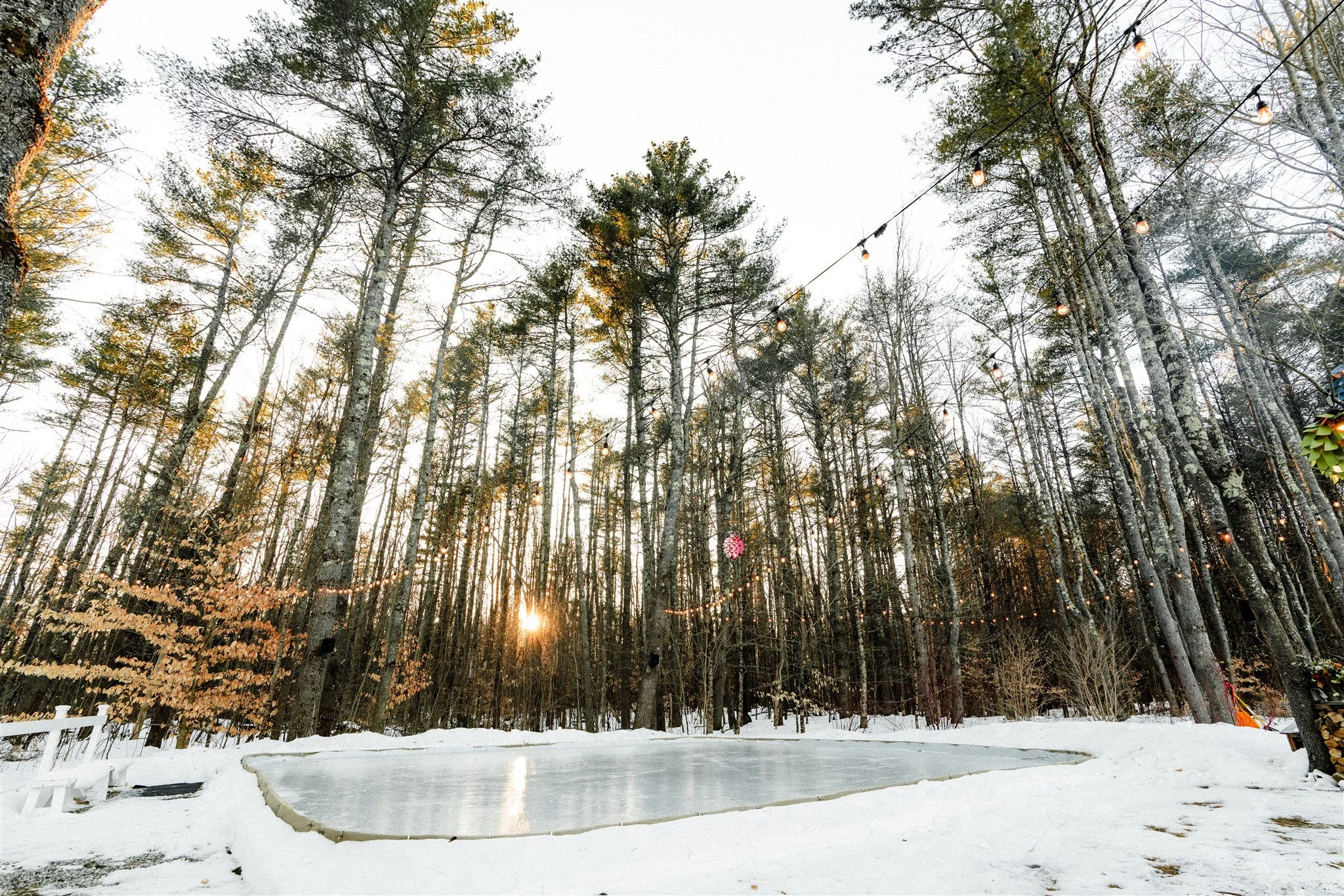 An ice skating rink surrounded by snow in a forested area, with string lights hanging between trees and a setting sun in the background.