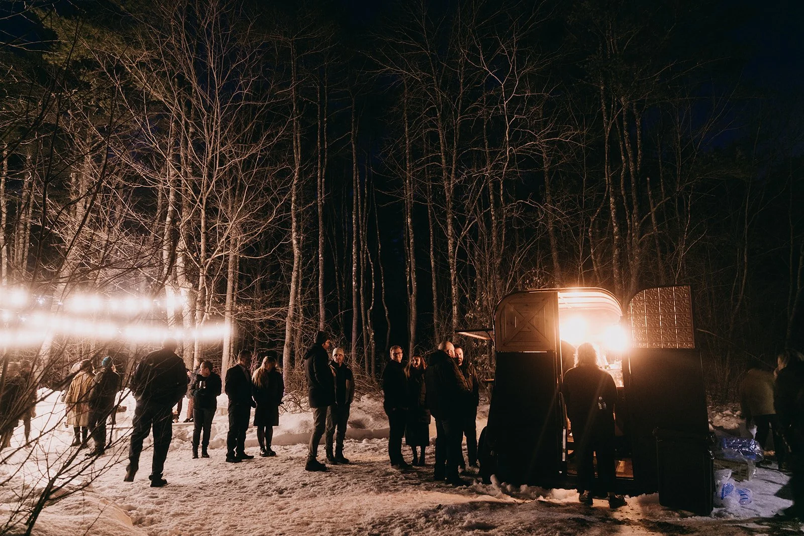 Group of people gathered outdoors in the snow at night, illuminated by a bright light source from a mobile food or drink truck, with a dense forest of leafless trees in the background.