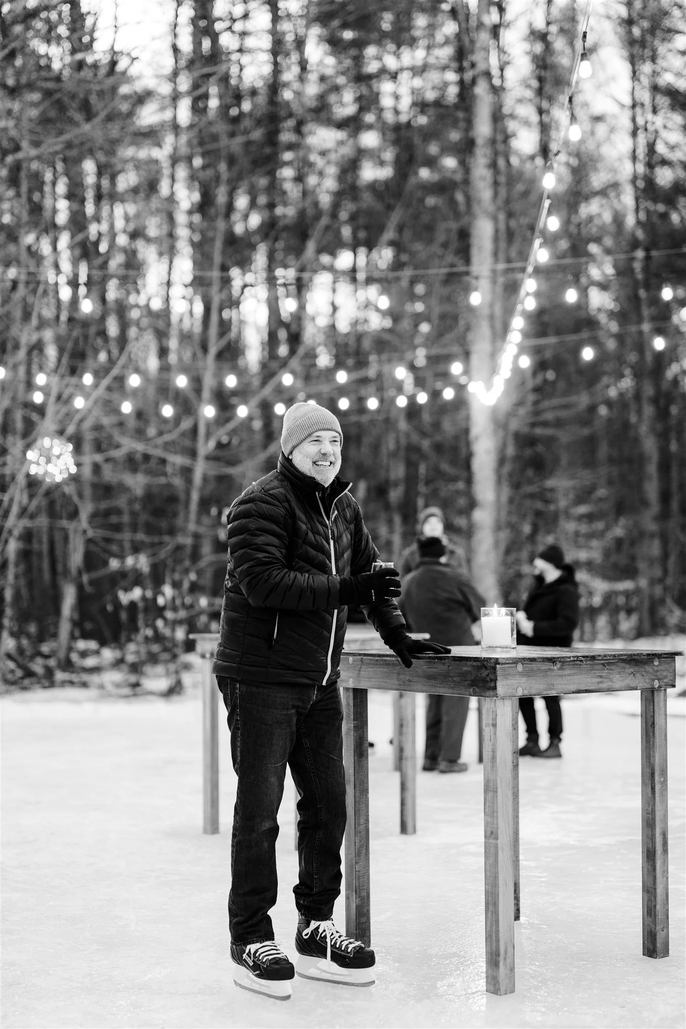 A man in a winter jacket, beanie, and gloves ice skating outdoors, smiling and holding a drink at a wooden table. In the background, two other people are talking near another table, with string lights hanging above in a wooded area.
