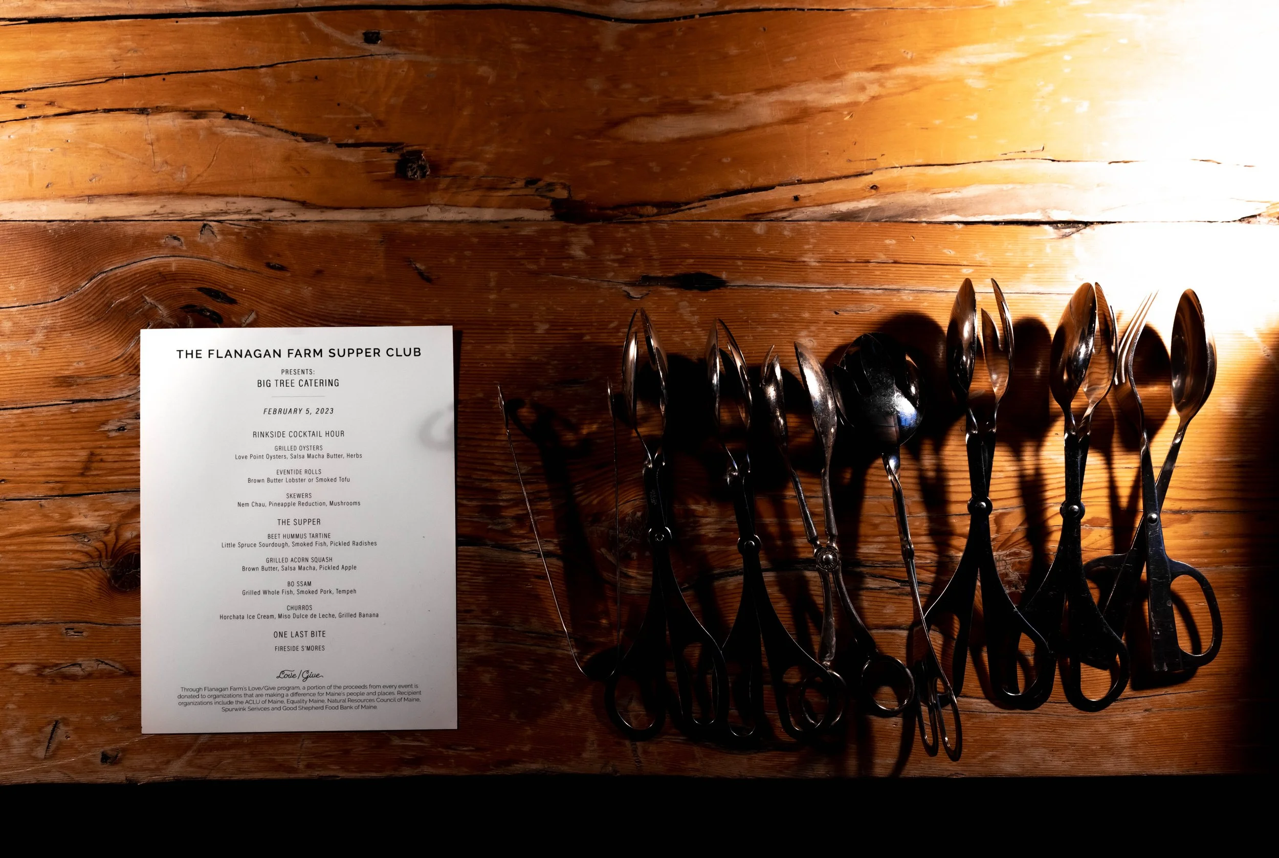 A printed menu and a row of black serving spoons on a wooden table at an event titled 'The Flanagan Farm Supper Club' with a scheduled date of February 5, 2023. The menu includes various dishes and desserts.