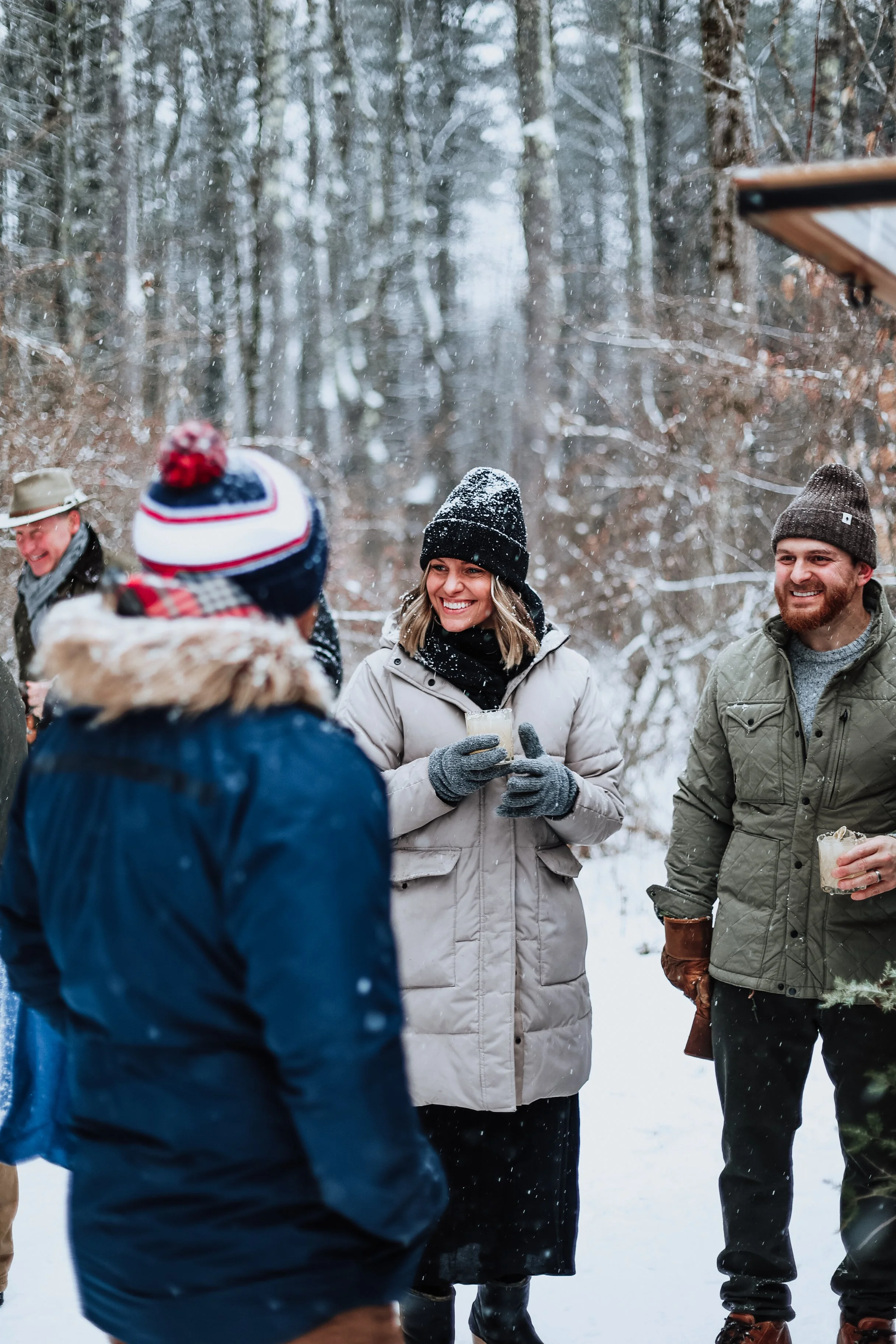 People outdoors in winter snow, smiling, chatting, with some holding drinks, dressed warmly in jackets, hats, and gloves.