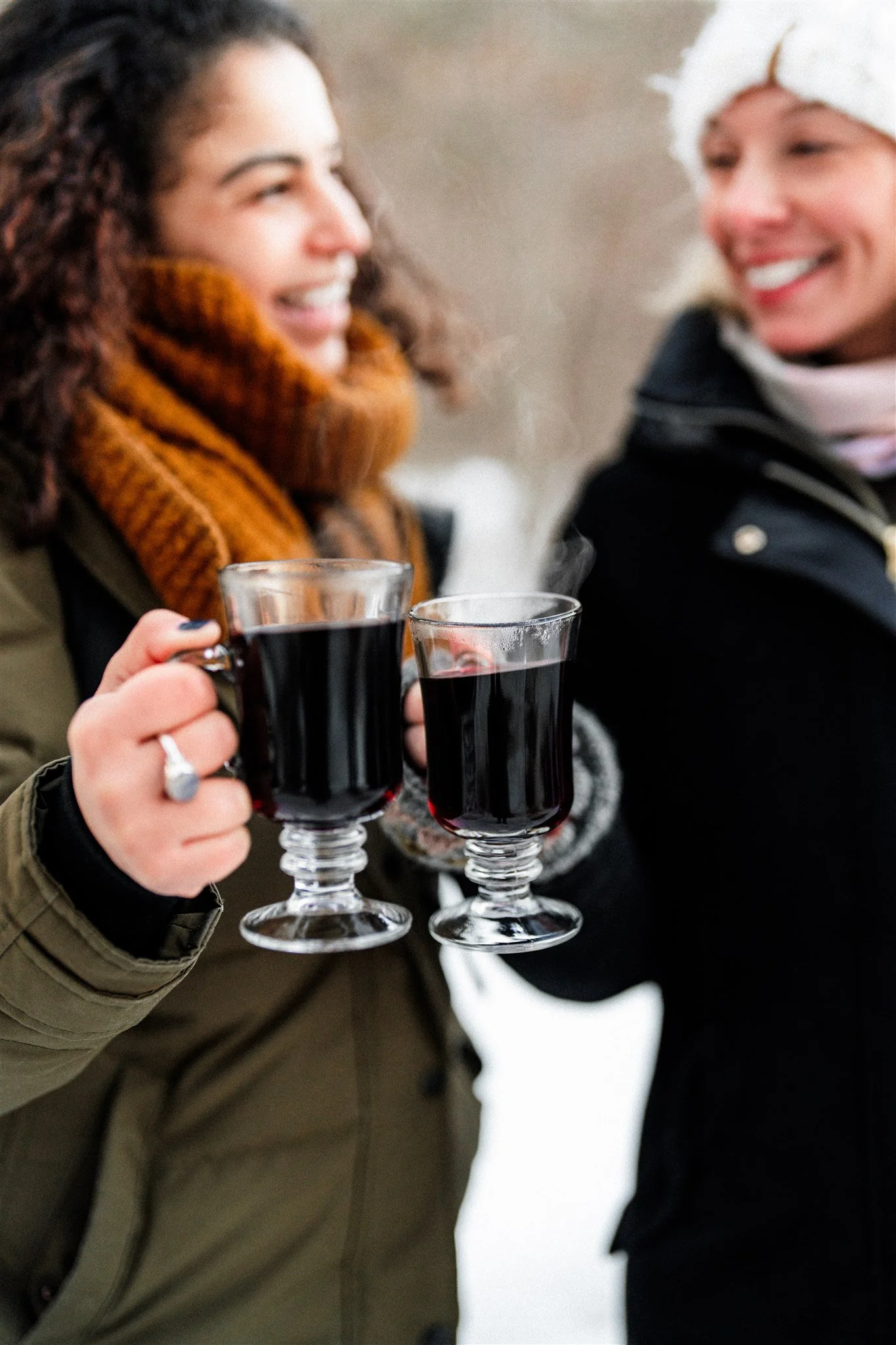 Two women smiling and holding glasses of hot dark beverage outdoors in winter, one wearing a brown scarf and the other a white hat.