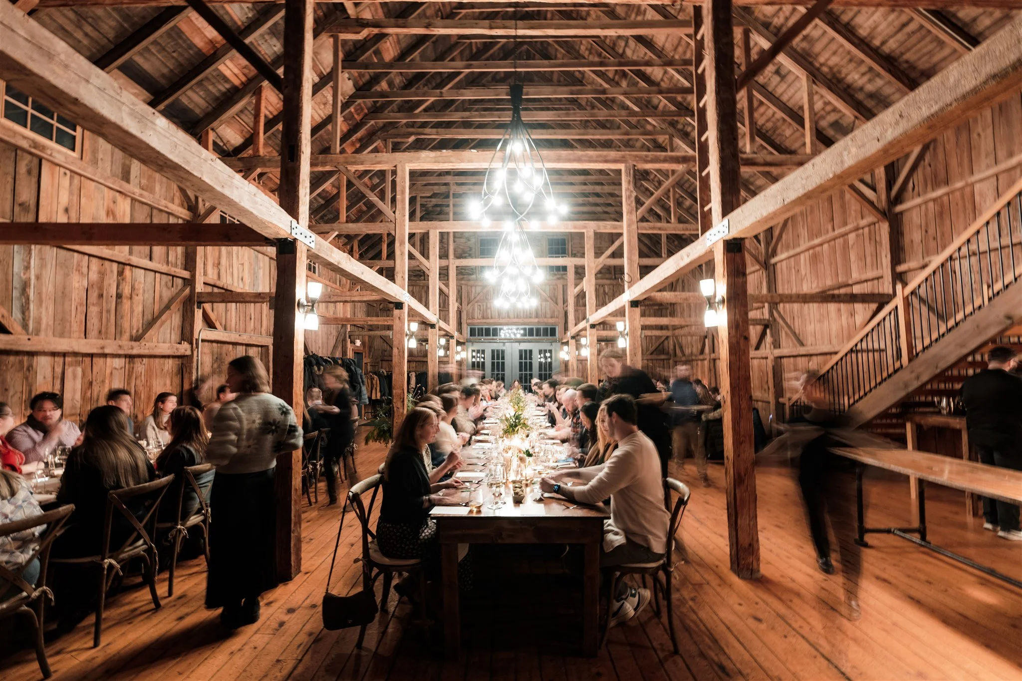 Brightly lit rustic wooden barn interior with a long dinner table and guests dining, chandeliers hanging from the ceiling, and a staircase on the right side.