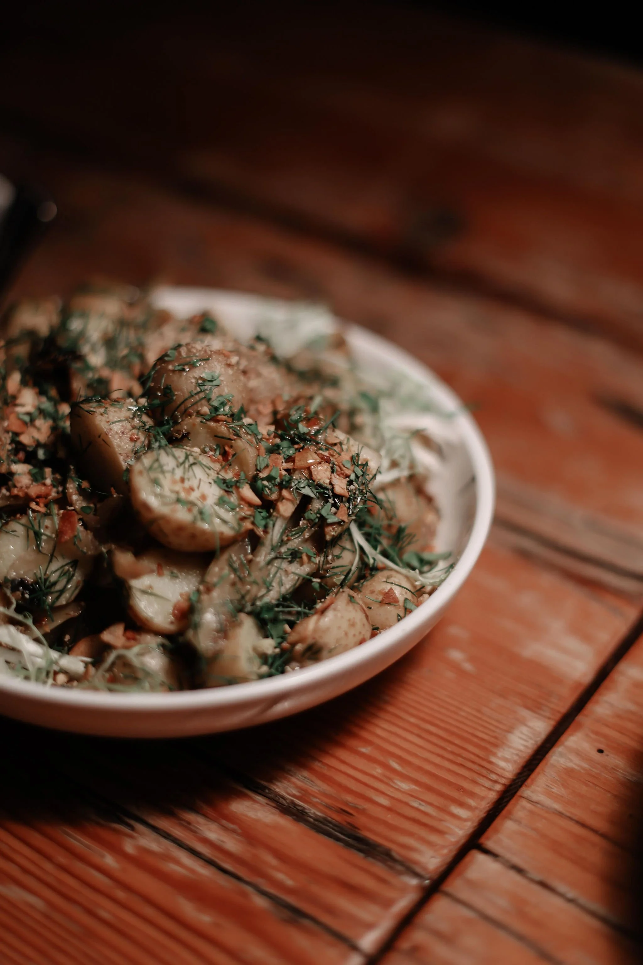 A white bowl filled with a salad containing sliced mushrooms, herbs, and possibly nuts, on a wooden table.