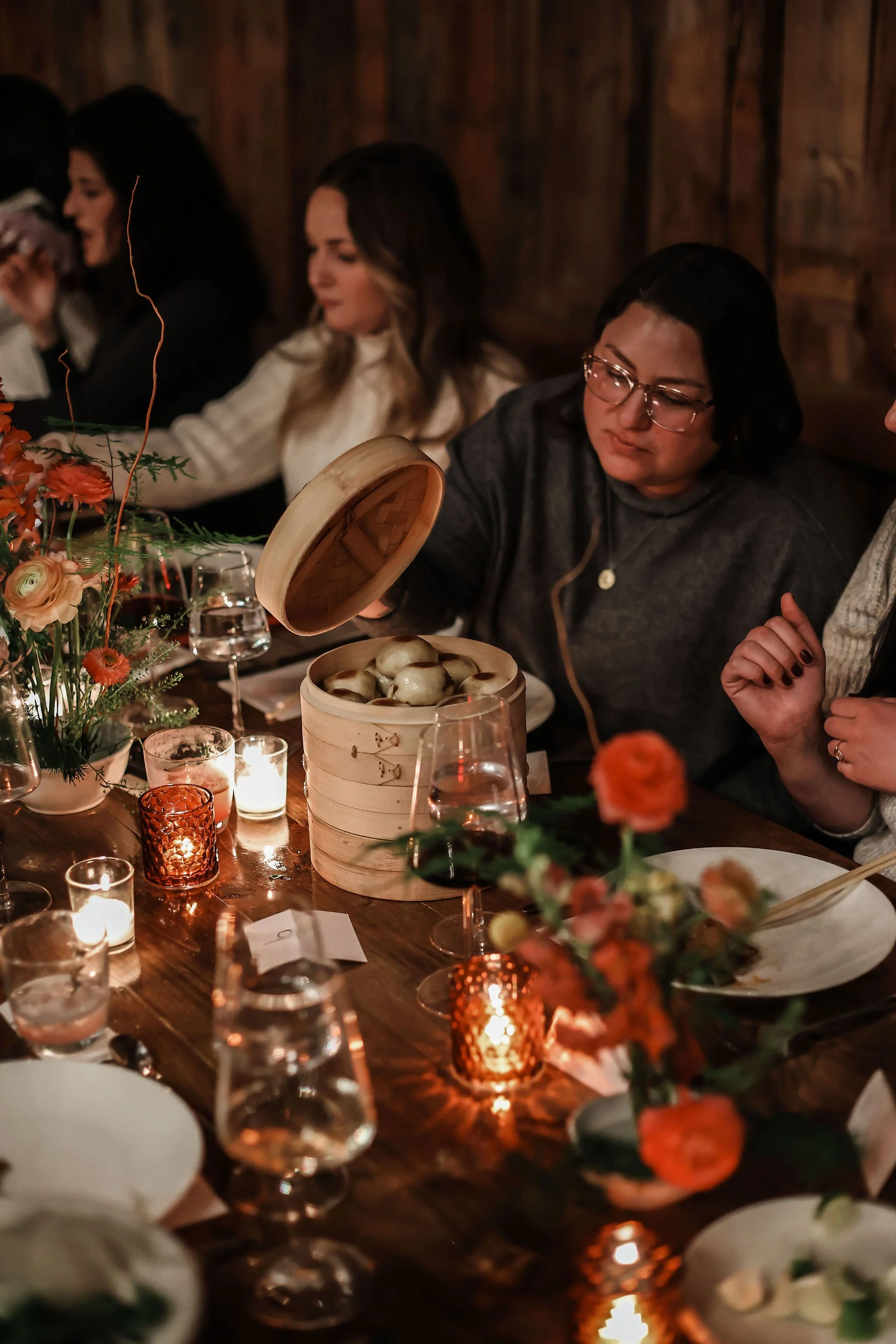 People gathered at a dinner table with candles and floral arrangements, sharing dim sum in a cozy, warmly lit setting.