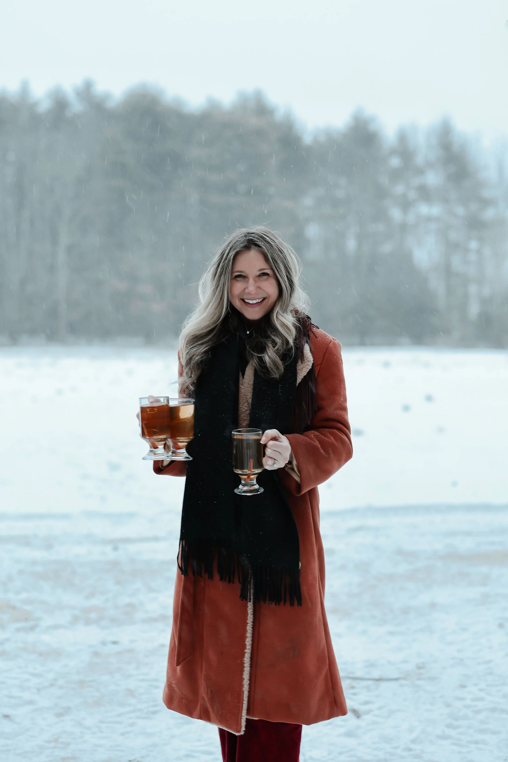 A smiling woman holding three glasses of drink outdoors in a snowy landscape.
