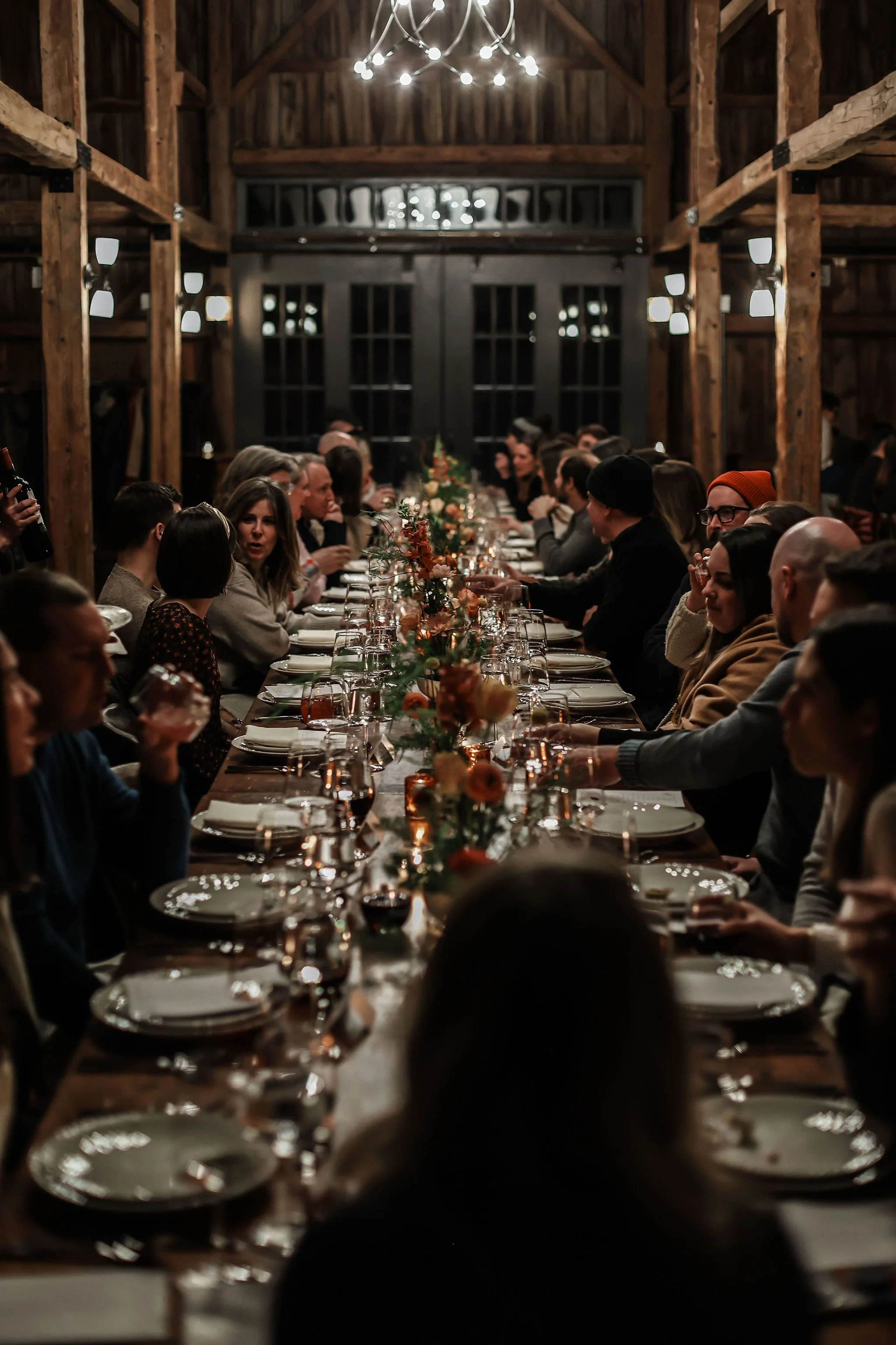 People gathered for a Christmas dinner party in a rustic barn with wooden beams and soft lighting, seated around a long table decorated with a floral centerpiece.