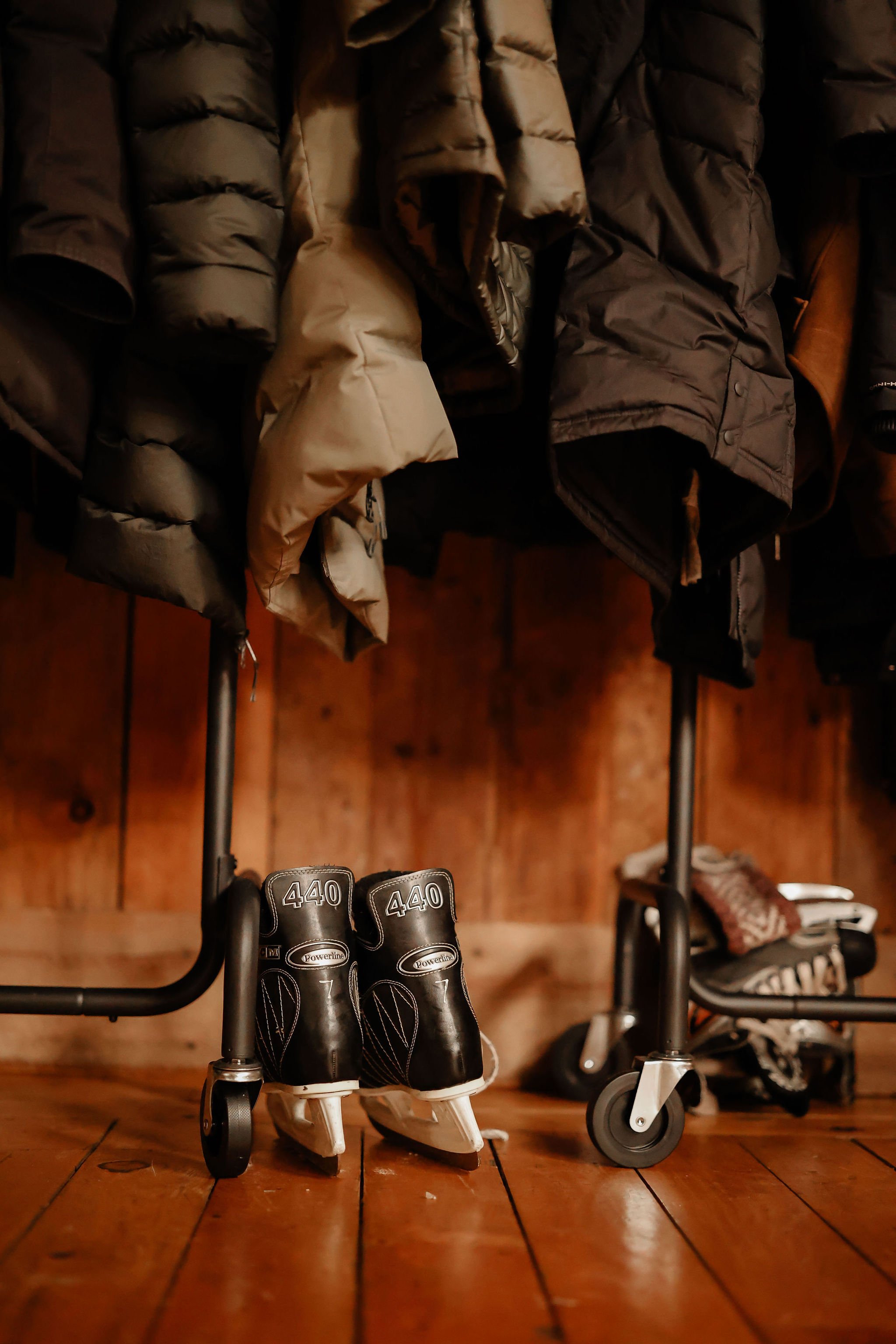 A pair of black ice hockey skates with the number 440 on them, placed on a wooden floor, below a clothing rack with winter jackets.